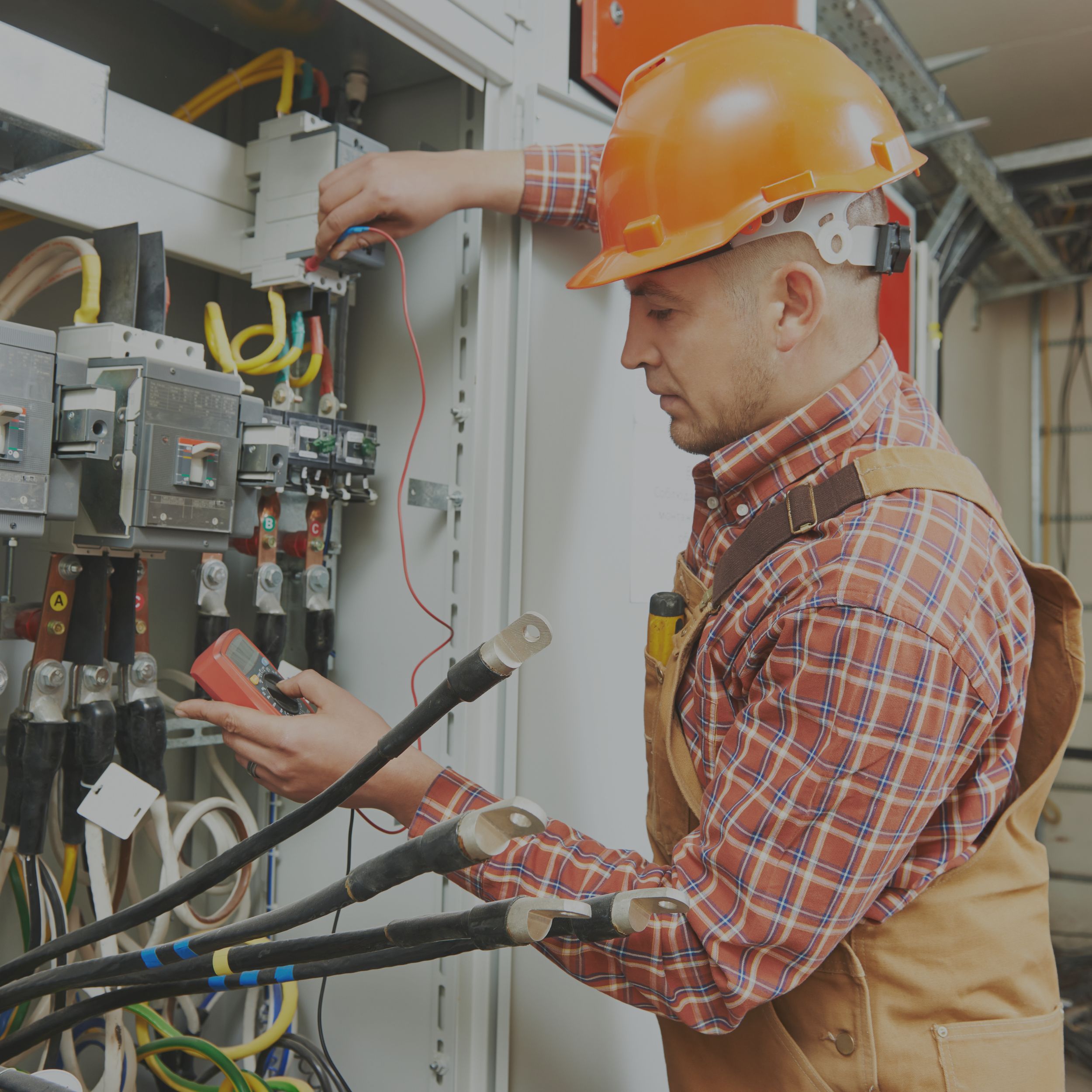 A technician wearing an orange hard hat and plaid shirt working inside an electrical control panel, using a multimeter to test the wiring.