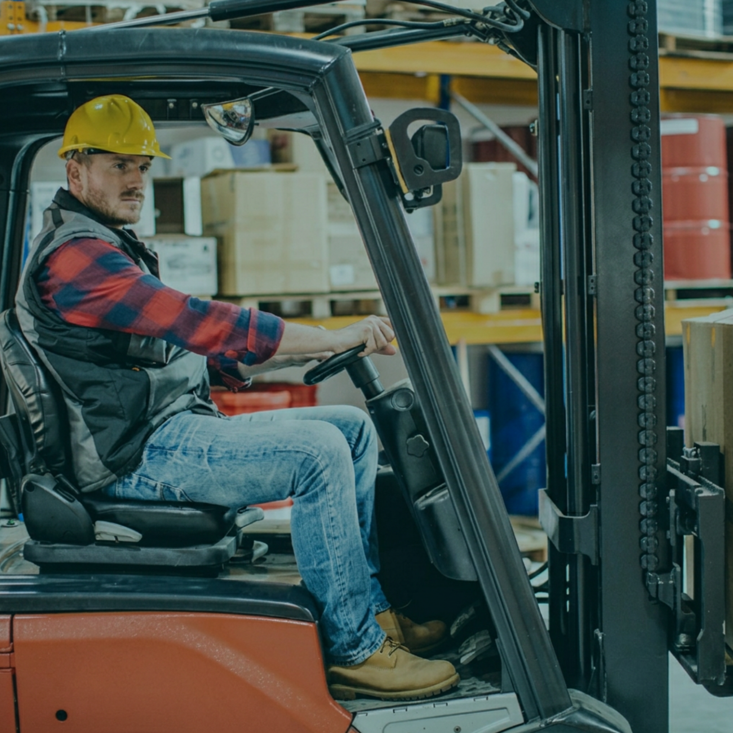 A man wearing a yellow safety helmet operating a forklift in a warehouse with shelves of boxes and supplies.
