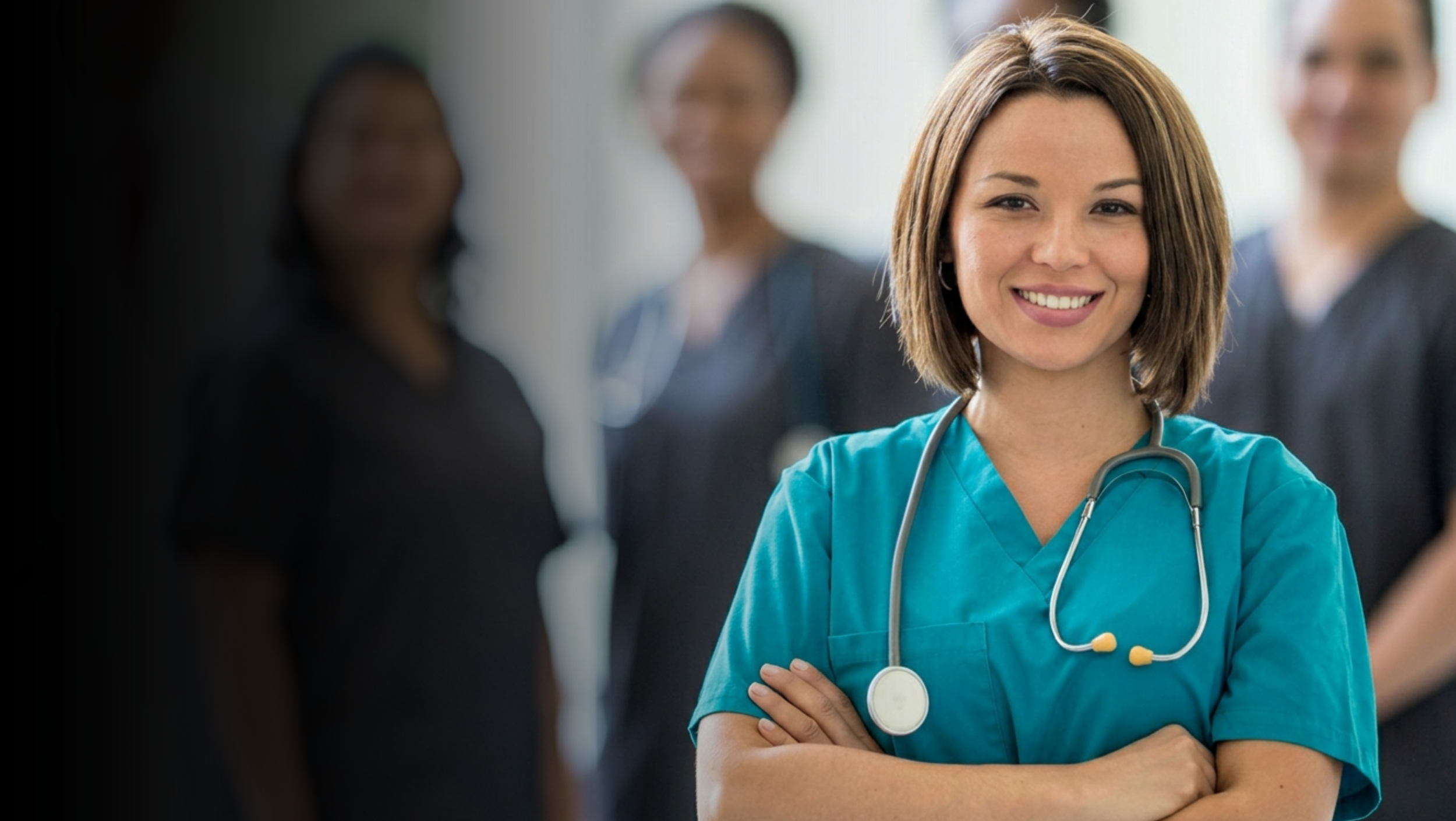 A smiling nurse with brown hair, wearing blue scrubs and a stethoscope around her neck, standing with arms crossed among her colleagues in a hospital or clinic setting.