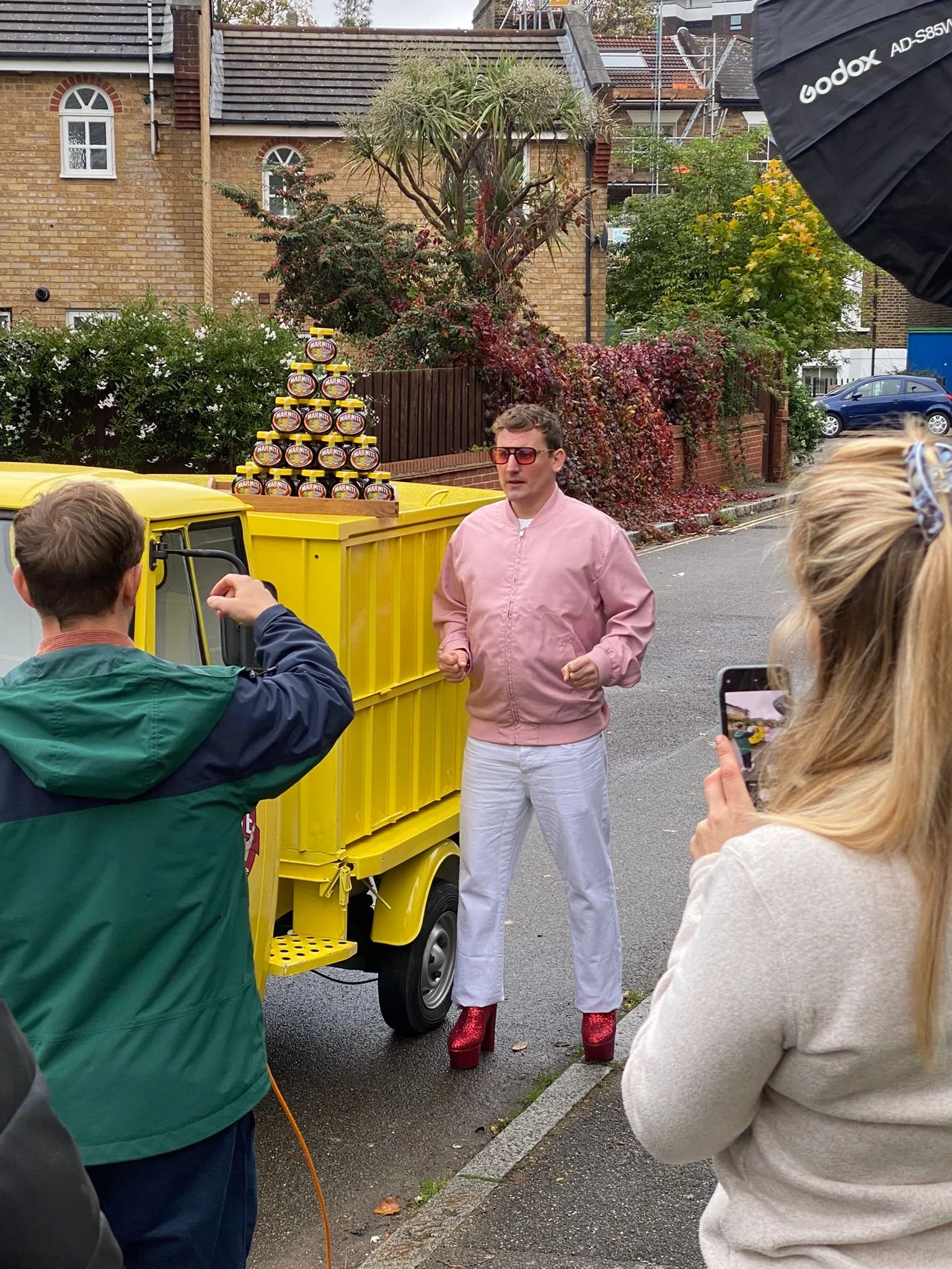 Man dressed in a pink jacket, white pants, and glittery red platform shoes standing next to a yellow cart with jars of marmalade stacked on top, as people take photos and record video around him, outdoors in a residential area.