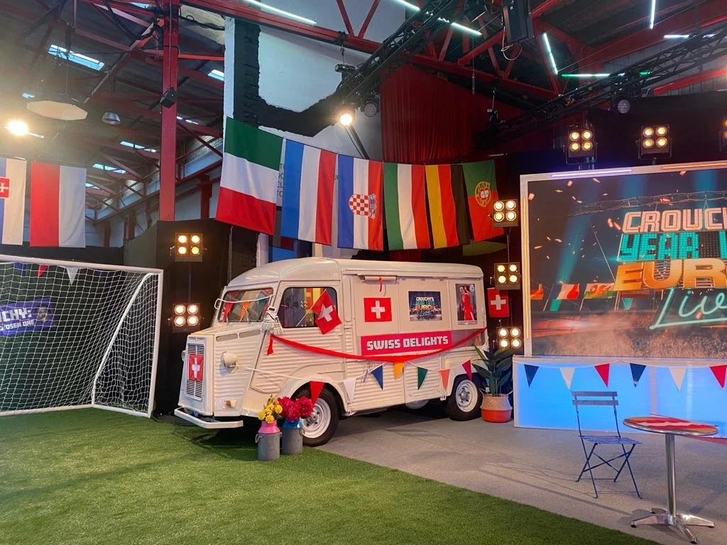 Decorative display with a vintage ice cream truck decorated with Swiss flags, positioned near a soccer goal. The background features a digital sign and European flags, with a festive atmosphere celebrating Europe.