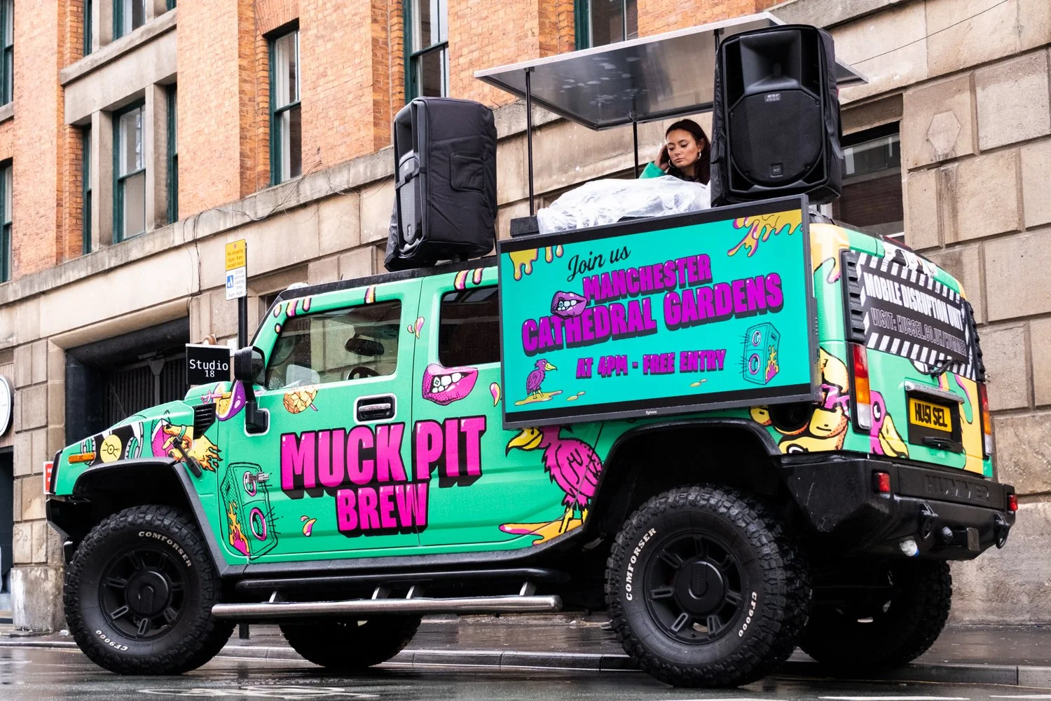 Decorated truck for Muck Pit Brew promoting an event at Manchester Cathedral Gardens with free entry at 4 PM, featuring vibrant graphics and a woman DJing on the roof.