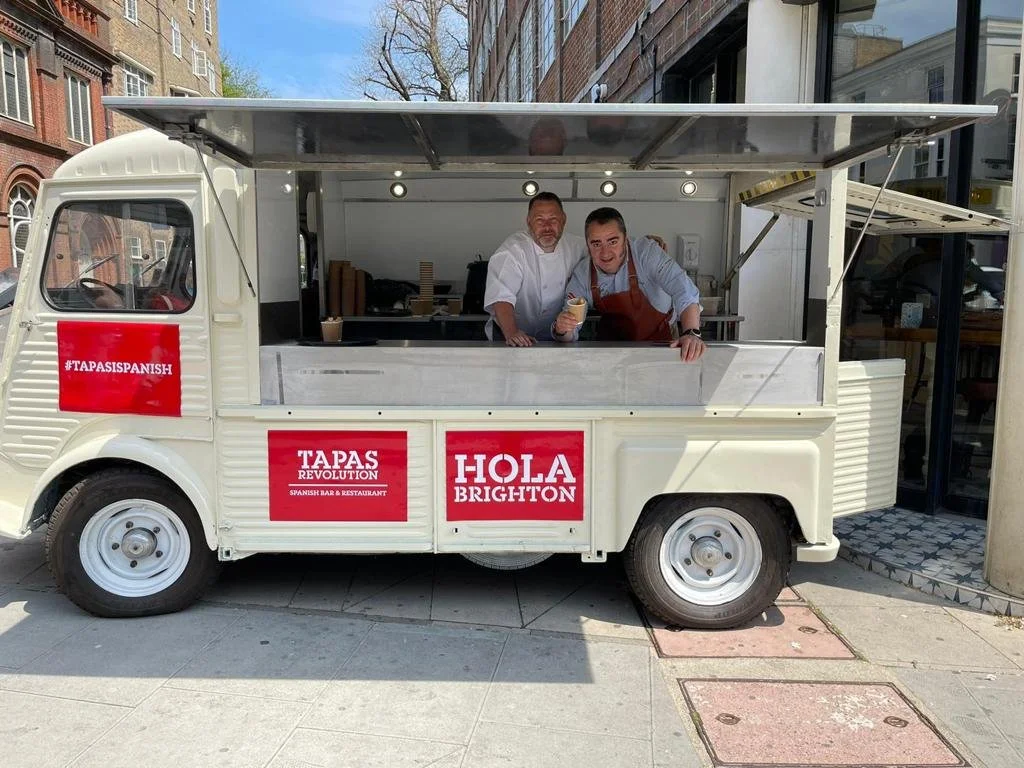 Two men are inside a small white food truck with red signs advertising Spanish food in Brighton, England. One man is wearing a white chef's coat and the other is wearing a light blue shirt with a red apron.