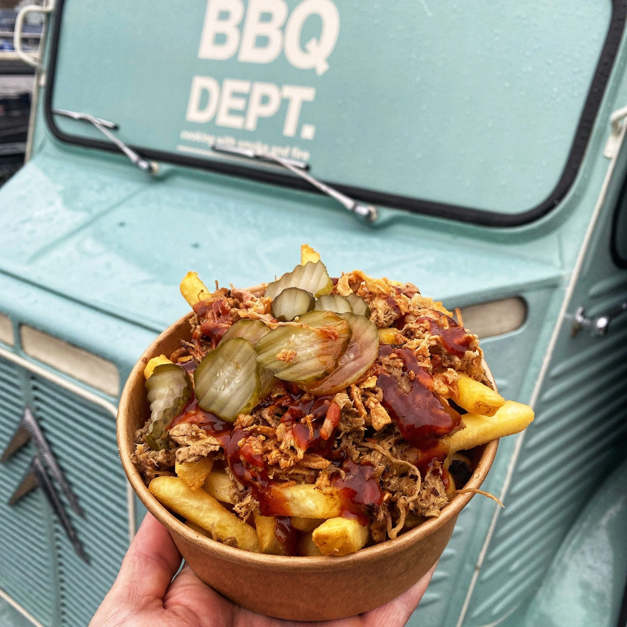 A hand holding a cup of loaded French fries topped with shredded meat, pickles, barbecue sauce, and other toppings, in front of a vintage Promovan H Van.