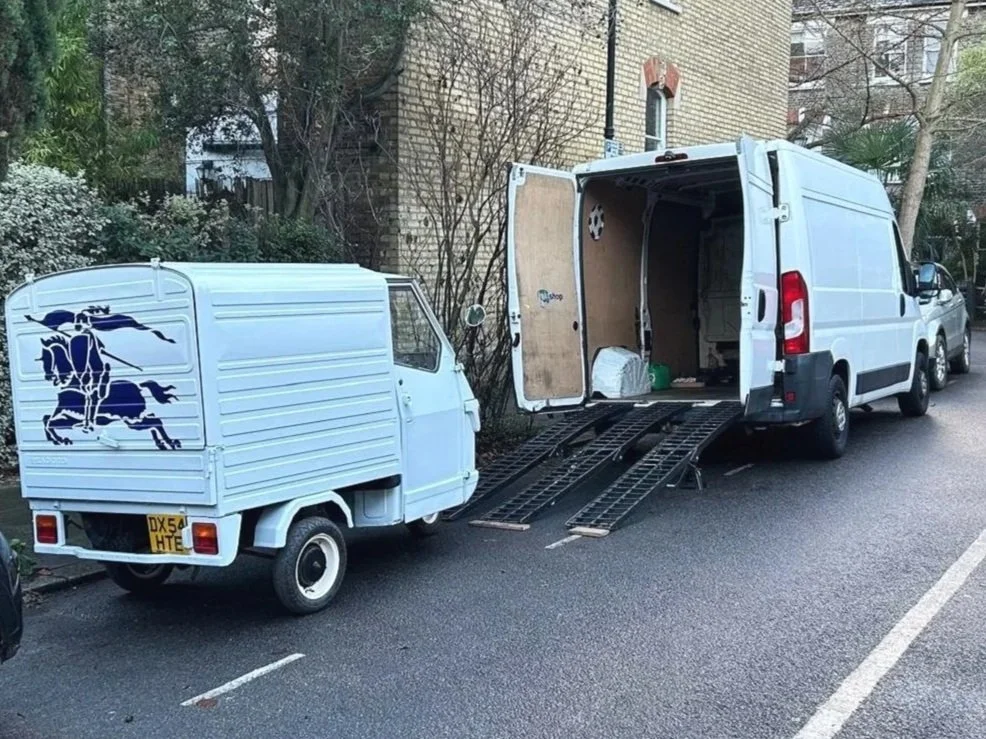 People crossing a street in front of a vintage food truck parked on the sidewalk, with a woman waiting near a street mailbox and a tree in the background.