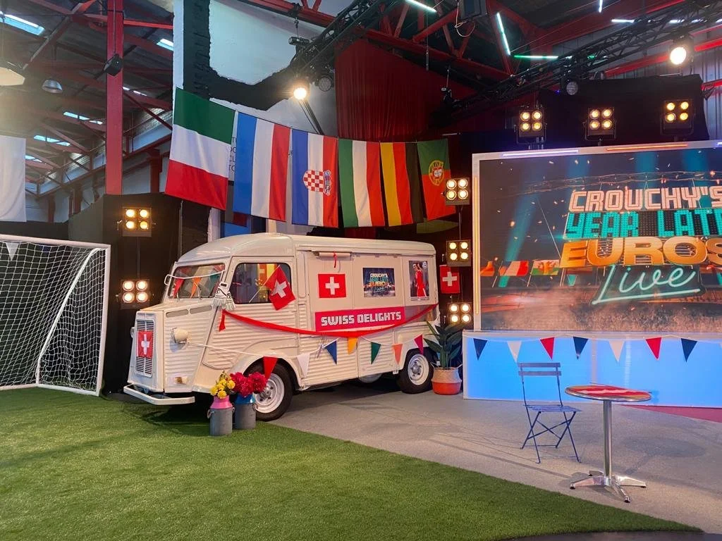 A vintage ice cream truck decorated with Swiss flags and flowers, outdoors at a TV studio or event space, with international flags hanging overhead and a large screen displaying 'Crouchy's Year Late Euros Live.'