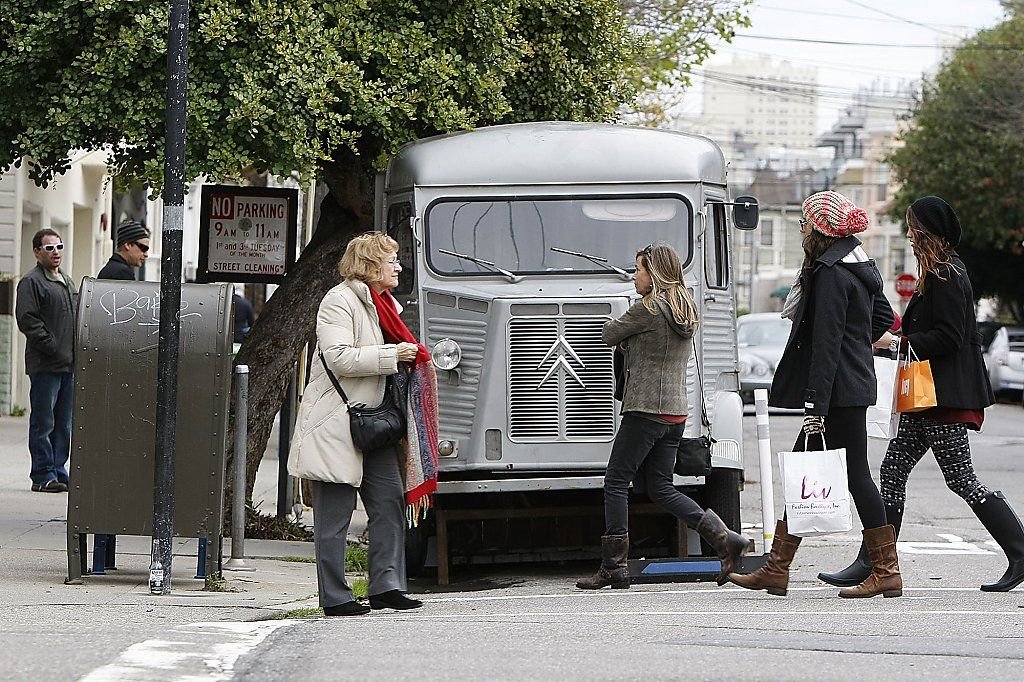 People crossing a street in front of a vintage food truck parked on the sidewalk, with a woman waiting near a street mailbox and a tree in the background.