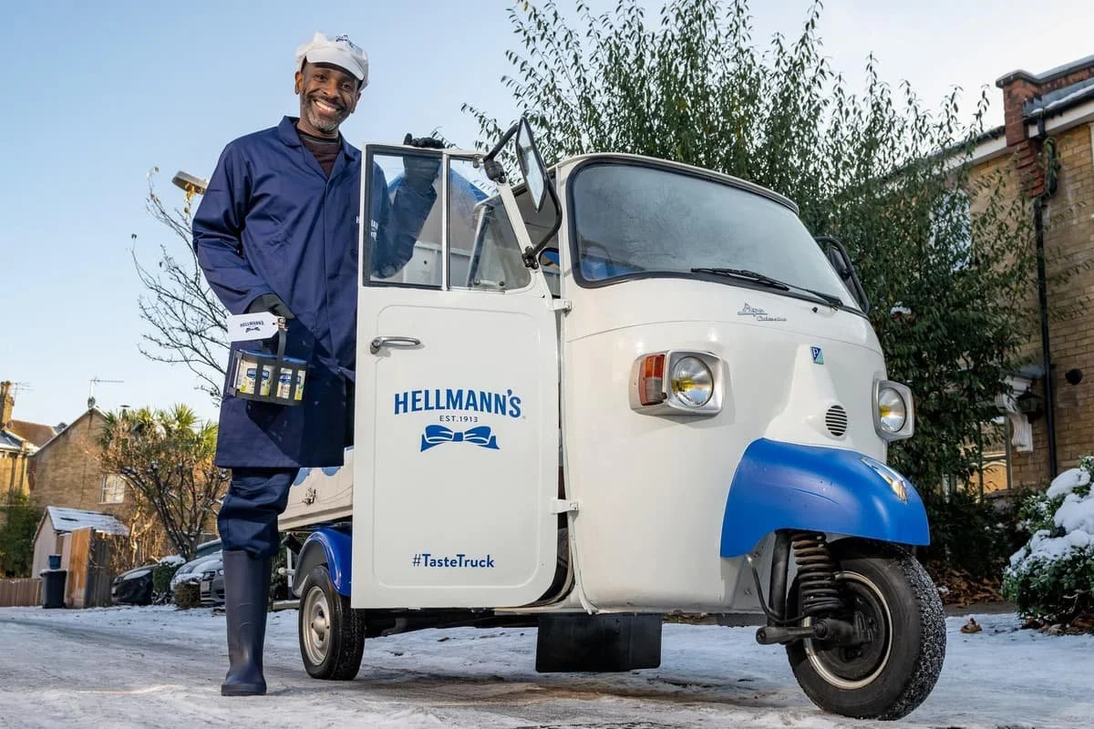 A man standing next to a small white and blue ice cream or food delivery tricycle, holding a box of bottled drinks, on a snowy street with houses and trees in the background.