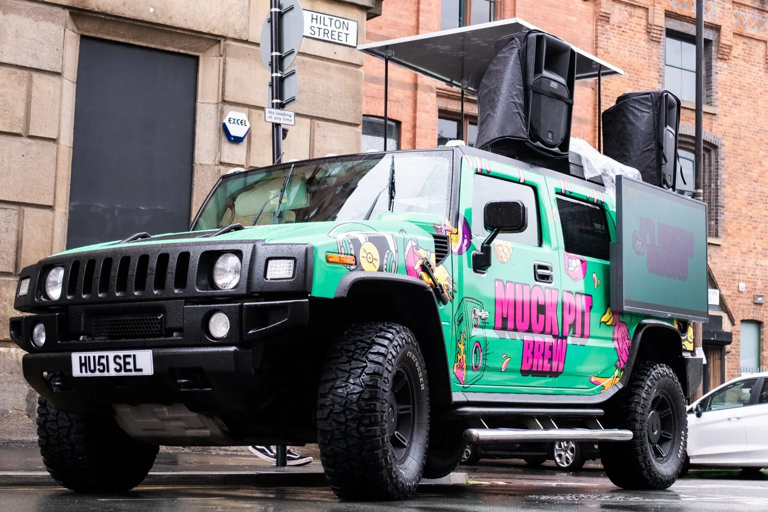 A colorful food truck with a graffiti-style design and the words 'Muck Pit Brew' on its side, parked on a city street with buildings and a street sign that reads 'Hilton Street'. The truck has large black speakers on top.