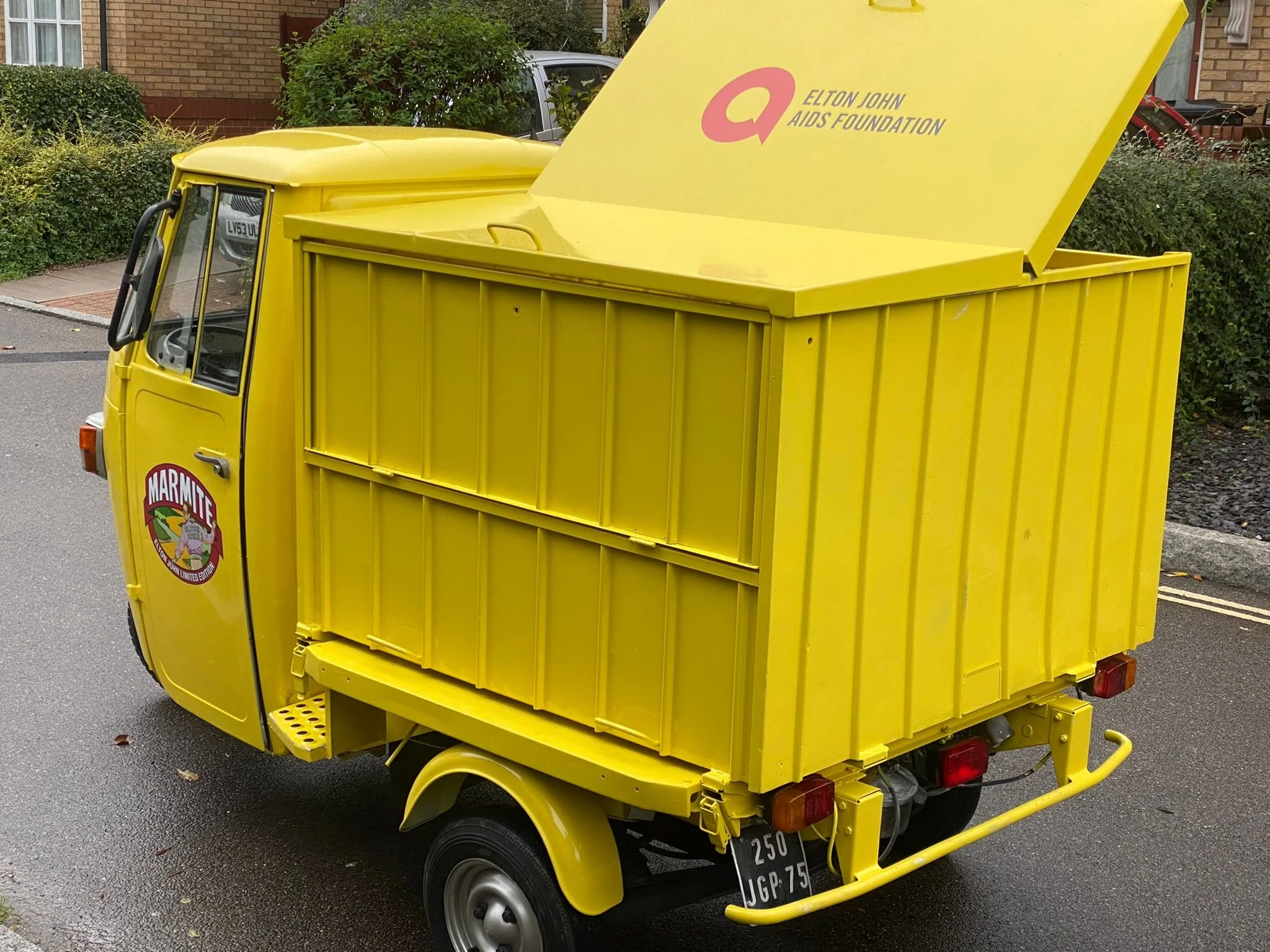 Yellow three-wheeled food truck with Marmite logo on the side and Elton John AIDS Foundation branding on the upper part of the truck's back, parked on a street with brick houses and greenery in the background.