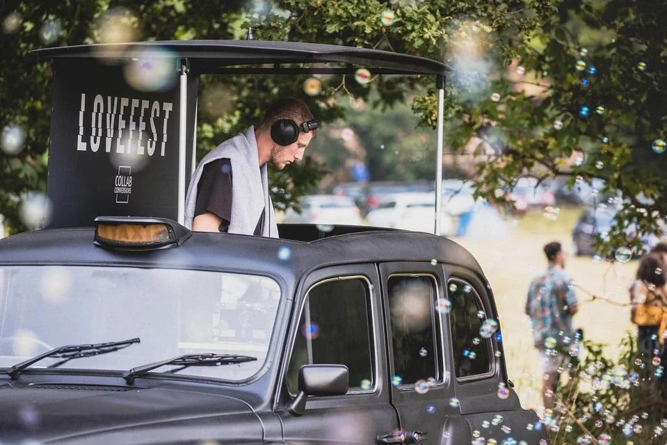 A DJ with headphones playing music from a booth on top of a vehicle at an outdoor event, with people in the background among trees and bubbles floating in the air.