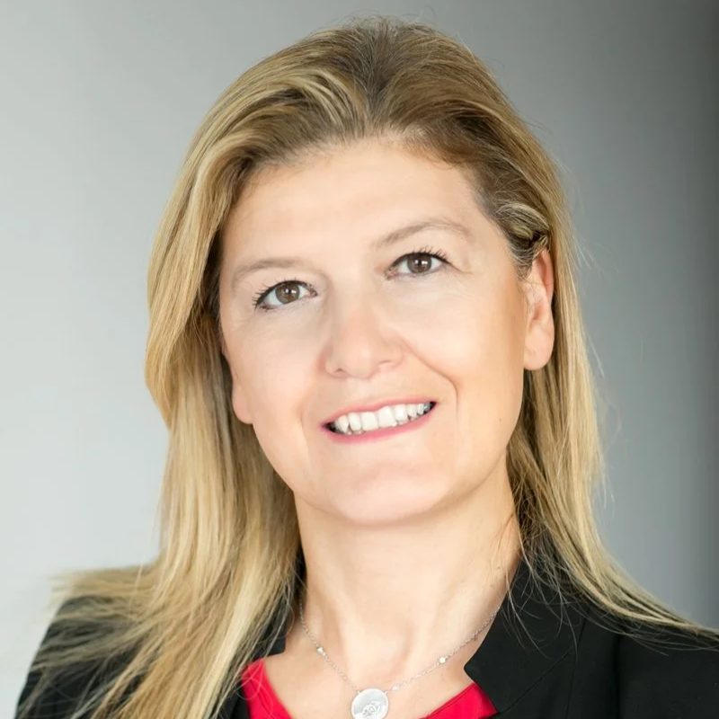 A professional woman with blonde hair, wearing a black blazer and red top, smiling at the camera against a plain gray background.