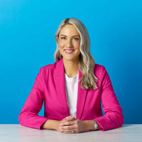 Portrait of a smiling woman with long blonde hair, wearing a pink blazer and white top, sitting at a table with a blue background.