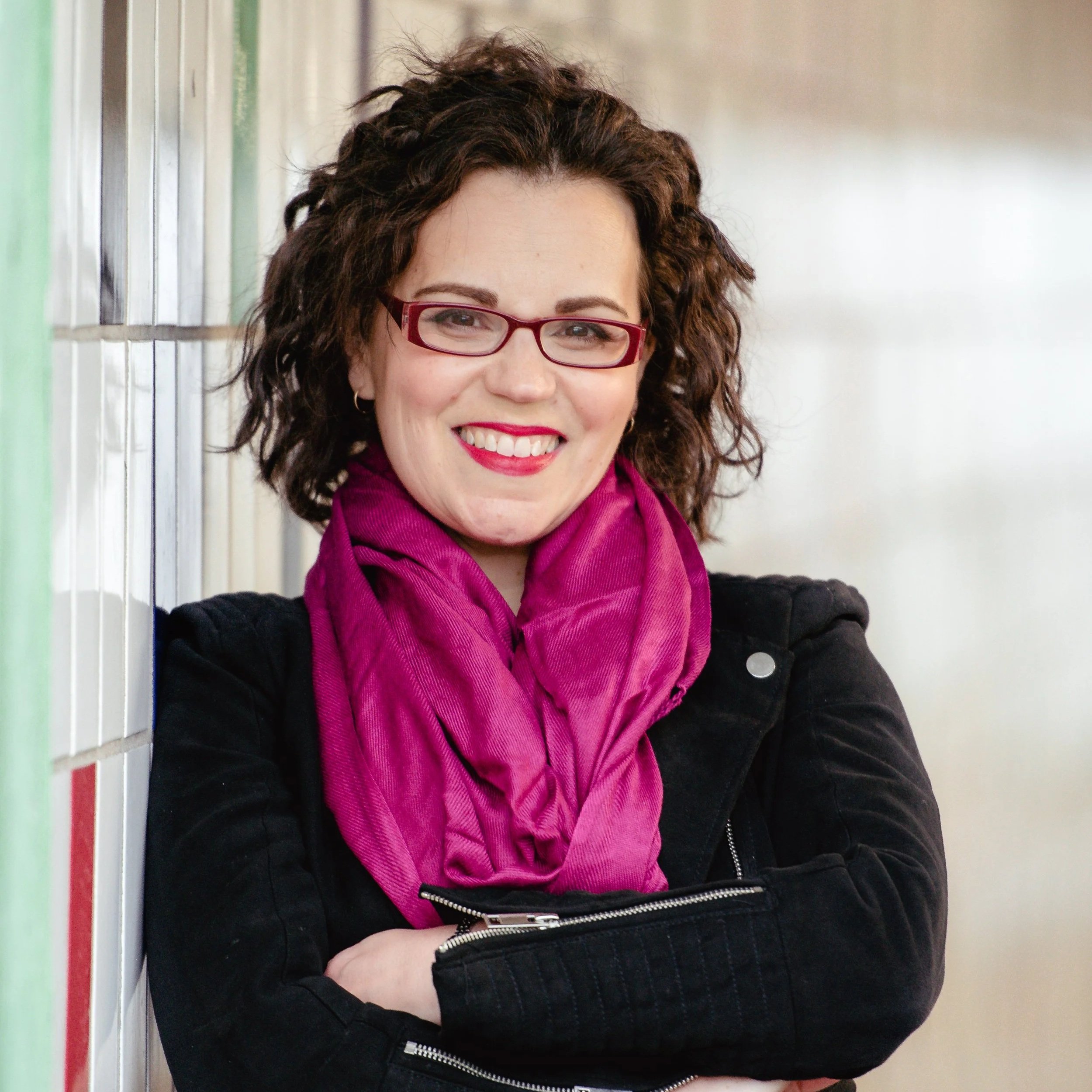A woman with curly brown hair, wearing glasses, a magenta scarf, and a black jacket, smiling with arms crossed.