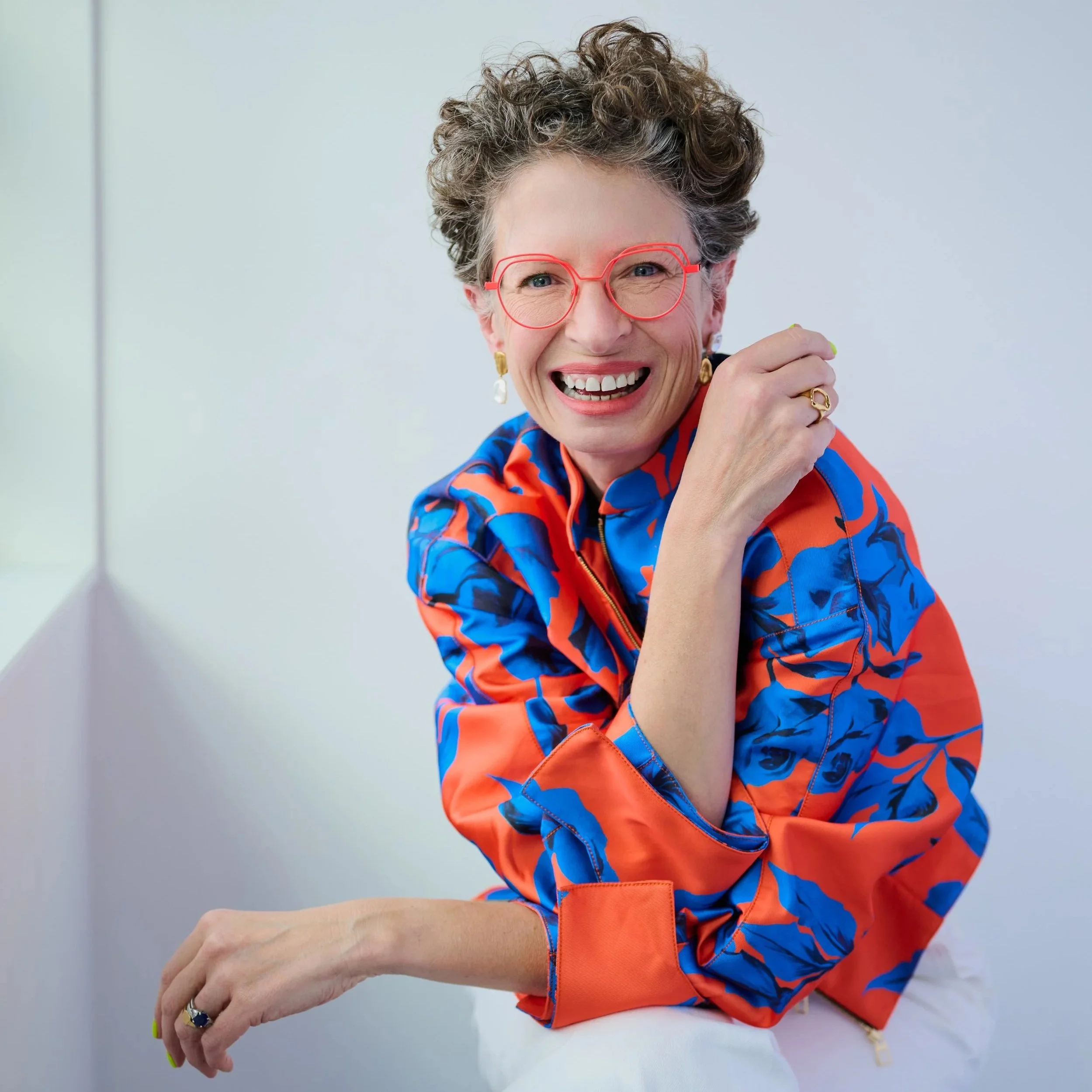 A smiling middle-aged woman with short curly gray hair, wearing red glasses, a colorful orange and blue patterned blouse, and gold earrings, is sitting and looking at the camera.