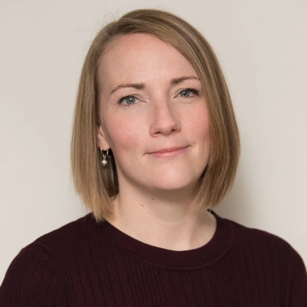 A woman with shoulder-length blonde hair, wearing a dark maroon sweater and earrings, standing against a plain light-colored background.