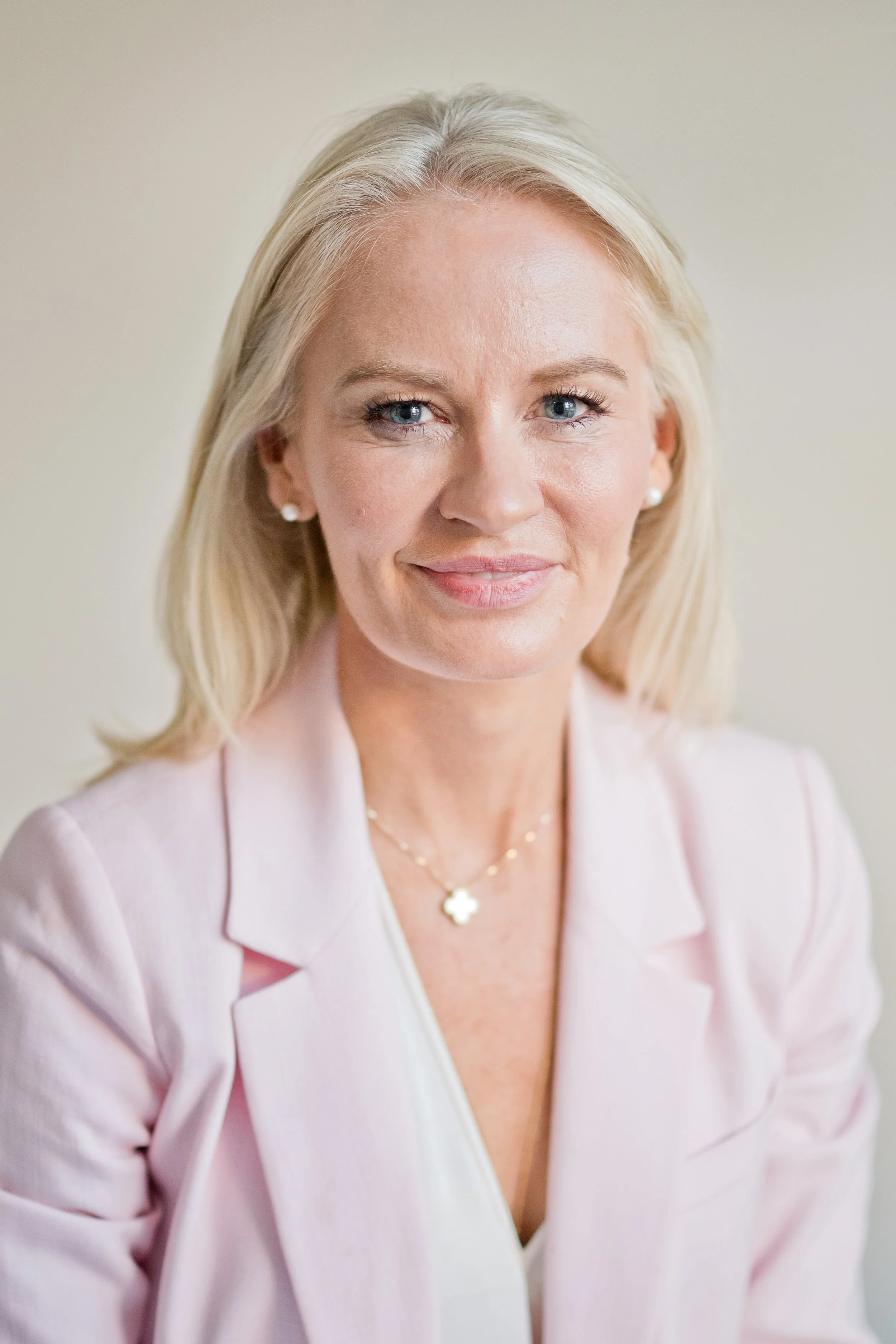A portrait of a blonde woman with blue eyes, wearing pearl earrings, a delicate beaded necklace with a four-leaf clover pendant, a light pink blazer, and a white blouse, smiling softly against a neutral background.