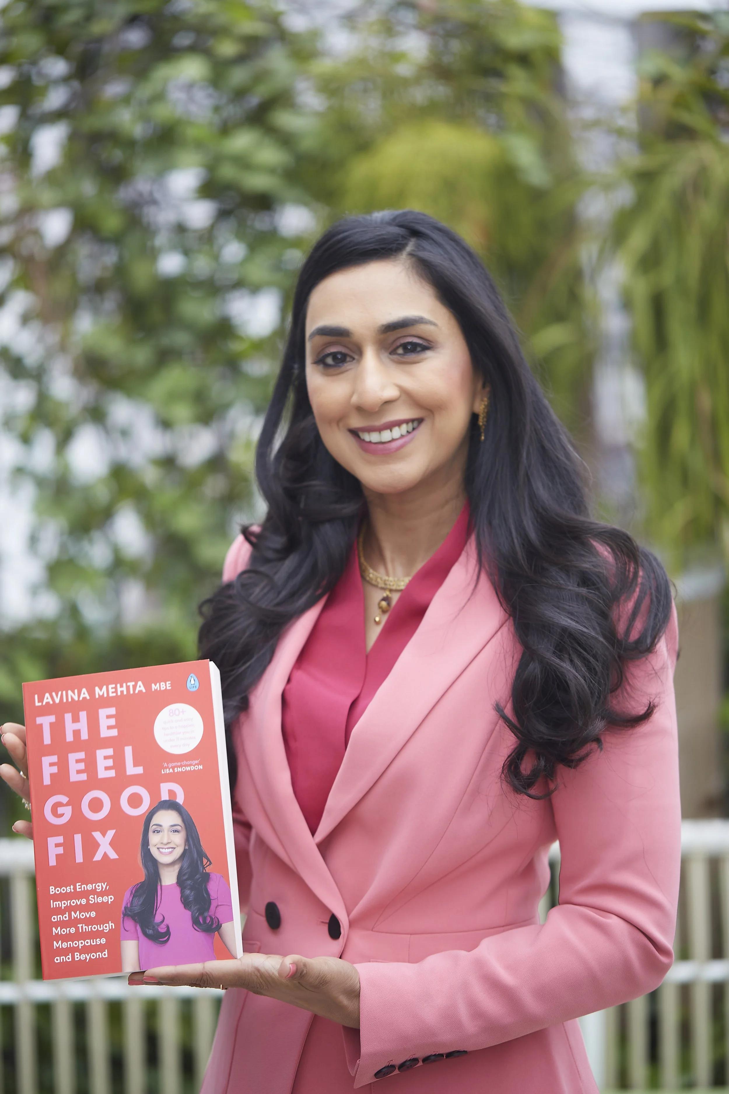 A smiling woman with long dark hair wearing a pink blazer and red top, holding a book titled 'The Feel Good Fix' by Lavina Mehta, outdoors with green foliage in the background.