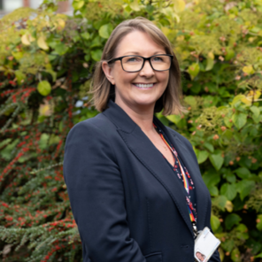 A woman with glasses and shoulder-length hair, smiling outdoors, standing in front of green and yellow foliage, wearing a navy blazer and a patterned blouse.