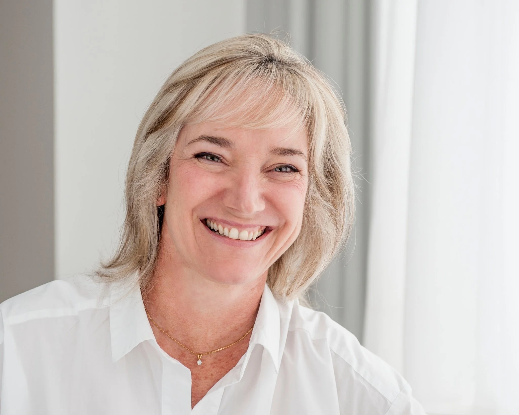 A smiling woman with blonde hair, wearing a white shirt, sitting in front of a window with sheer curtains.