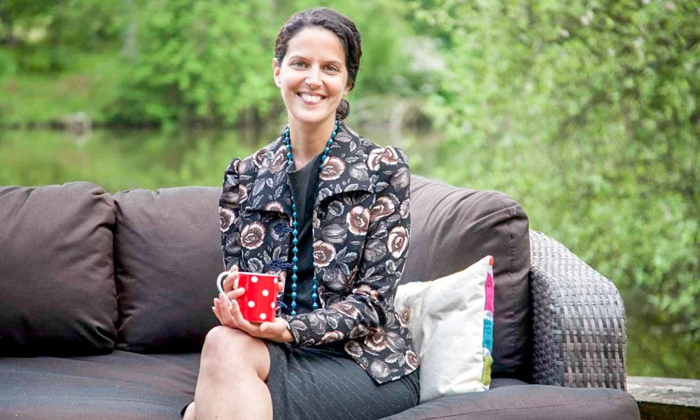 A woman sitting outdoors on a brown wicker couch, holding a red and white polka dot mug, smiling, with trees and a water body in the background.