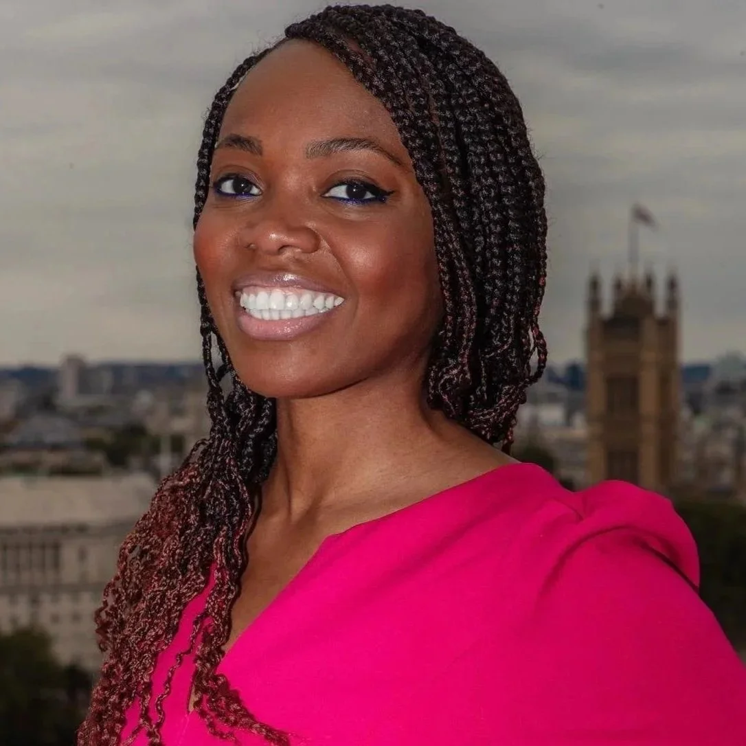 A woman smiling with a cityscape in the background, wearing a pink top and with braided hair.
