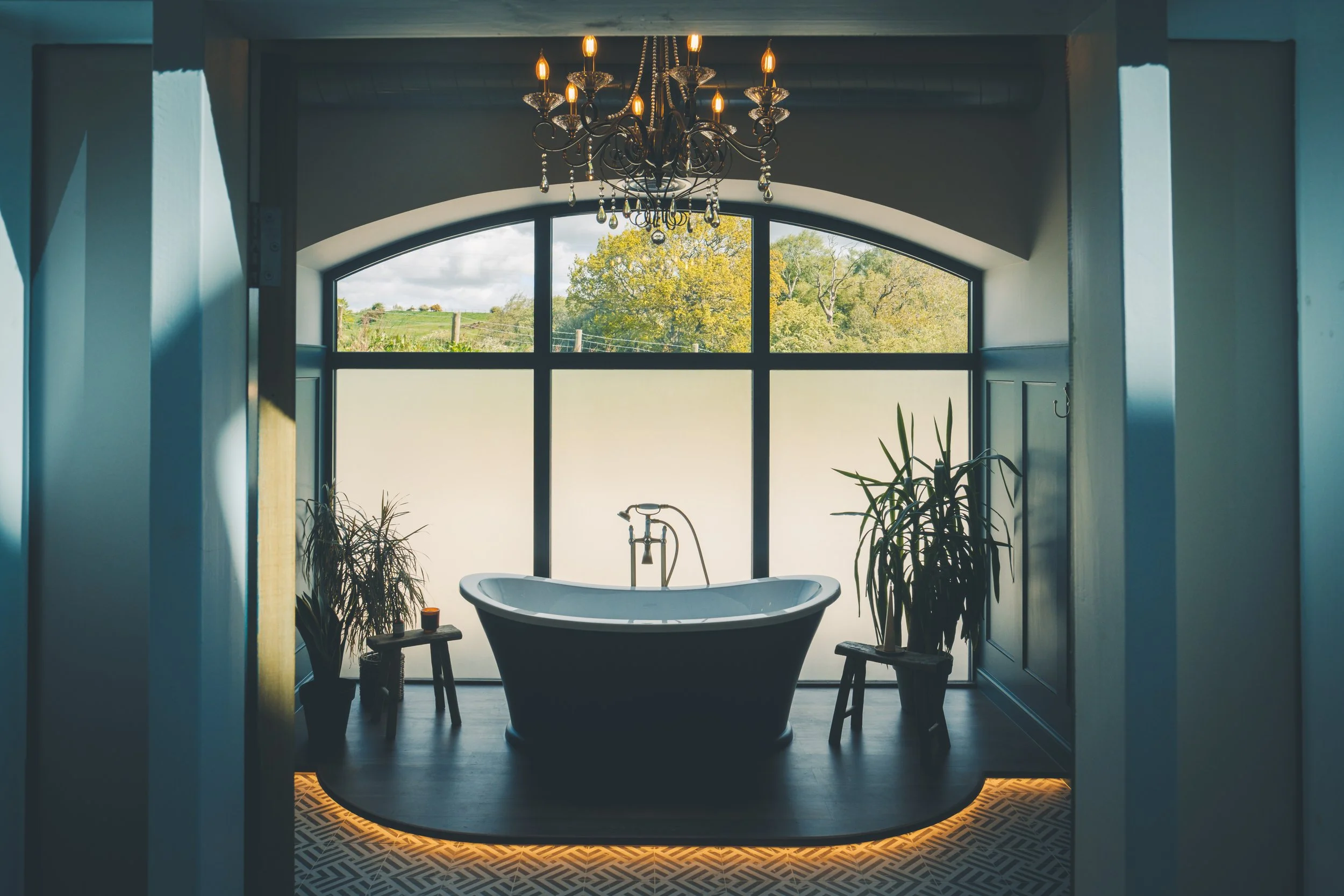 Bathroom with freestanding bathtub, chandeliers, large window showing trees outside, indoor plants, and patterned floor.