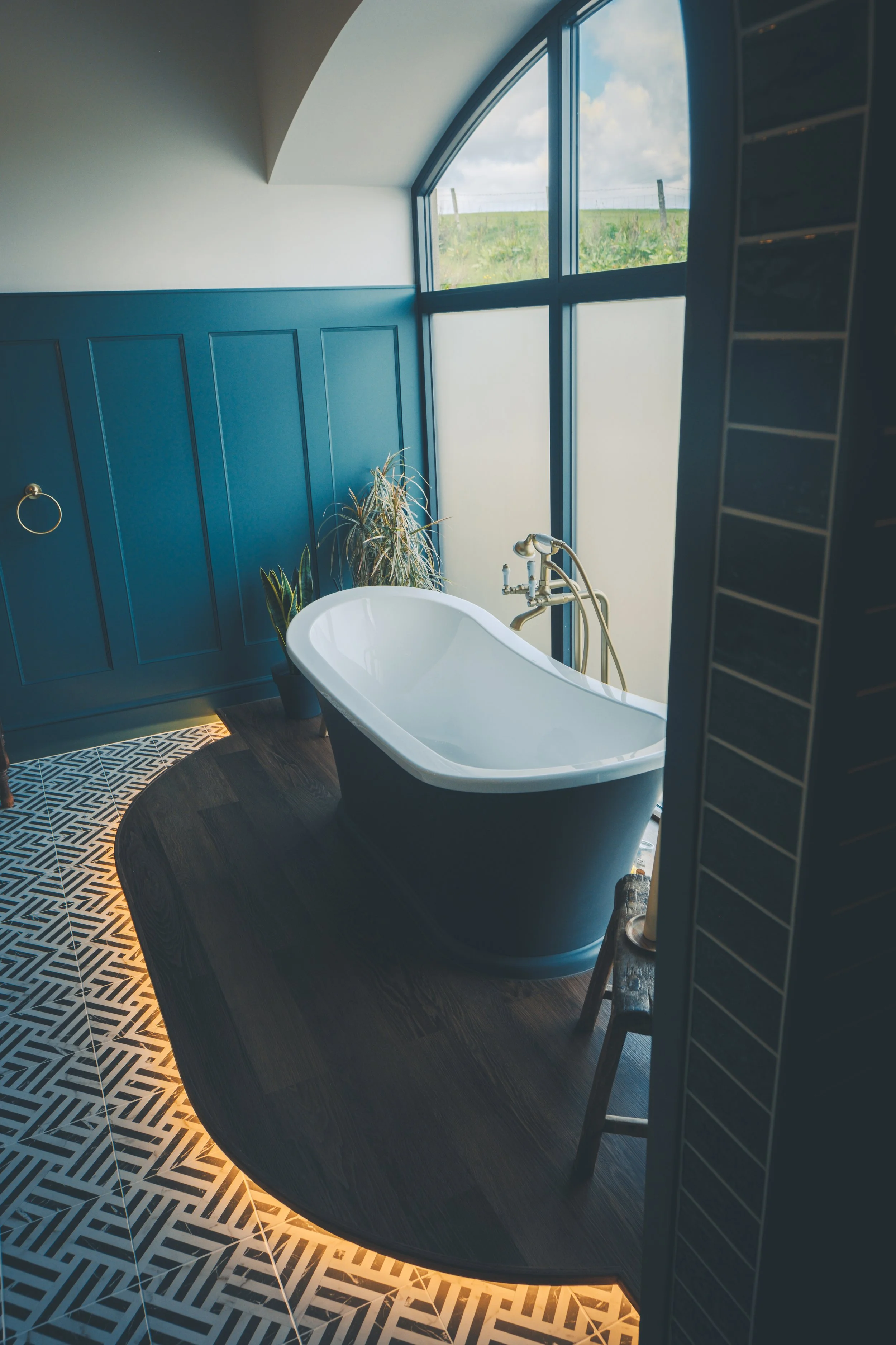 A bathroom with a white freestanding bathtub on dark wooden flooring next to a blue paneled wall and a large window showing an outside landscape with green grass and a cloudy sky. There are some green plants beside the bathtub and a table holding a l