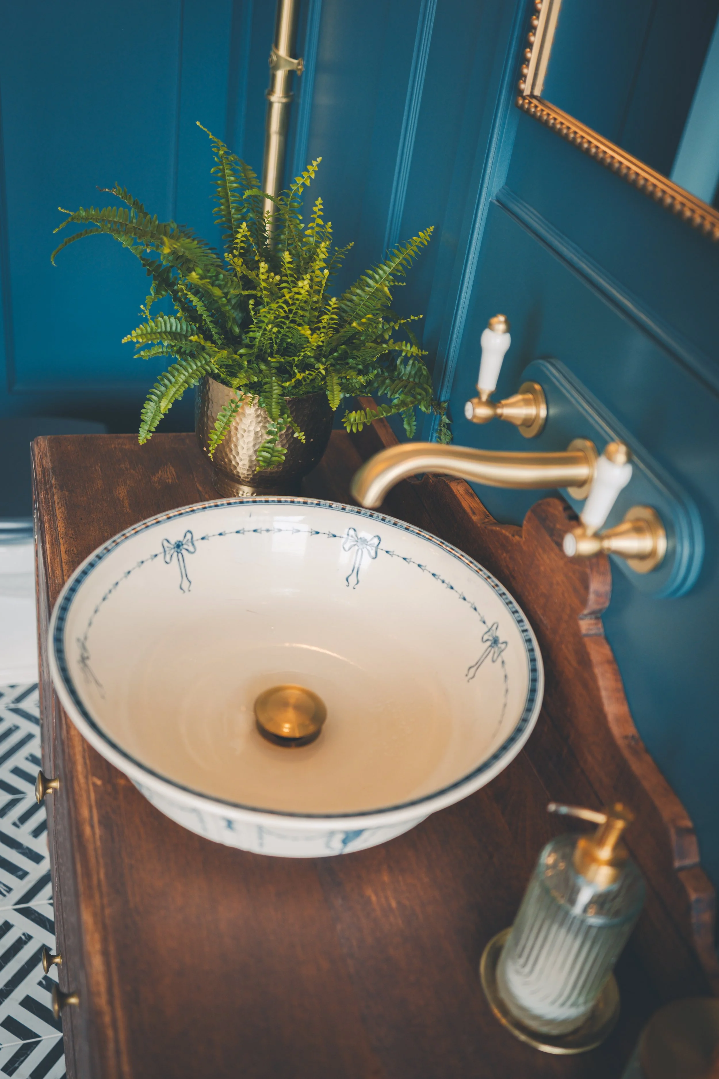 Close-up view of a decorative bathroom sink with a ceramic basin featuring blue bows, a gold drain, and a brass faucet. A green potted fern in a hammered metal pot is placed on a wooden vanity against a teal wall with a gold mirror frame.