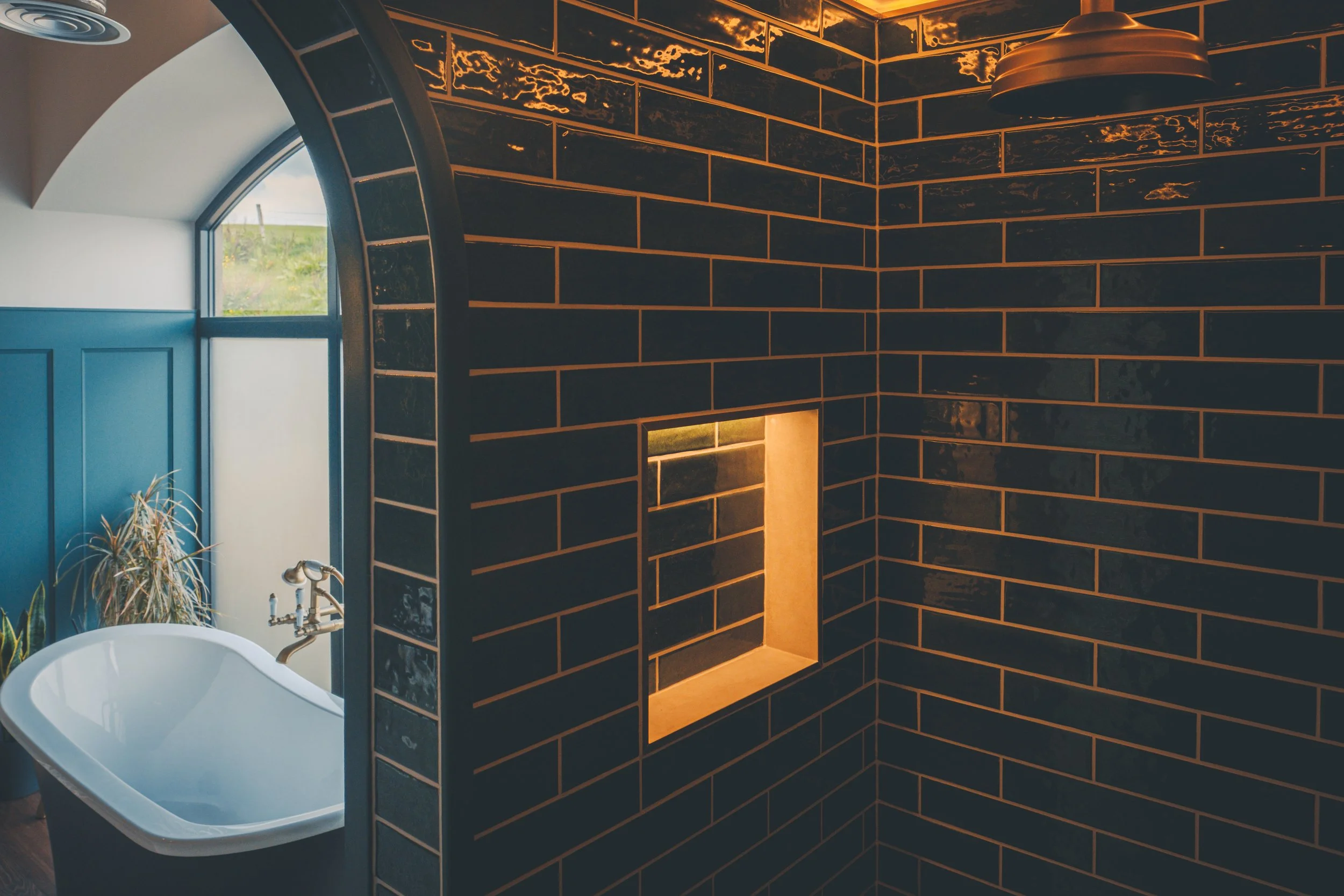 A modern bathroom with dark brick-patterned tiles on the walls, a square opening with backlighting, a small window showing green grass outside, a white bathtub with a curved edge, and a tall potted plant near the bathtub.