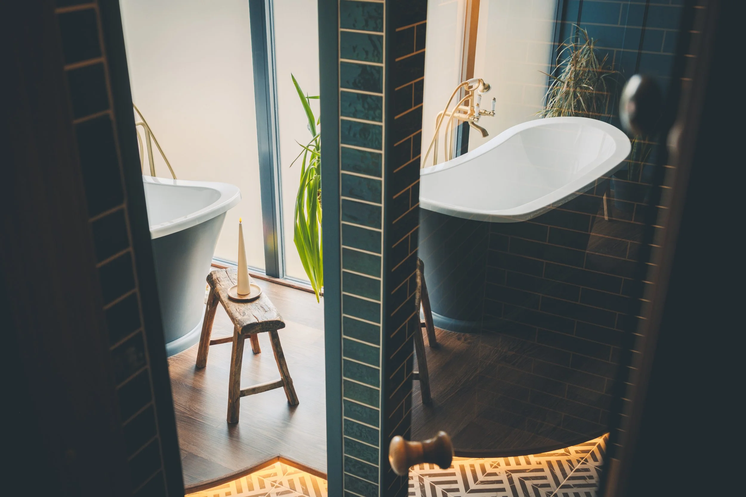 View of a bathroom with a white bathtub next to a window with plants outside, a vintage wooden stool, a brass faucet, dark tiled walls, and patterned floor tiles, seen through a partially opened door.