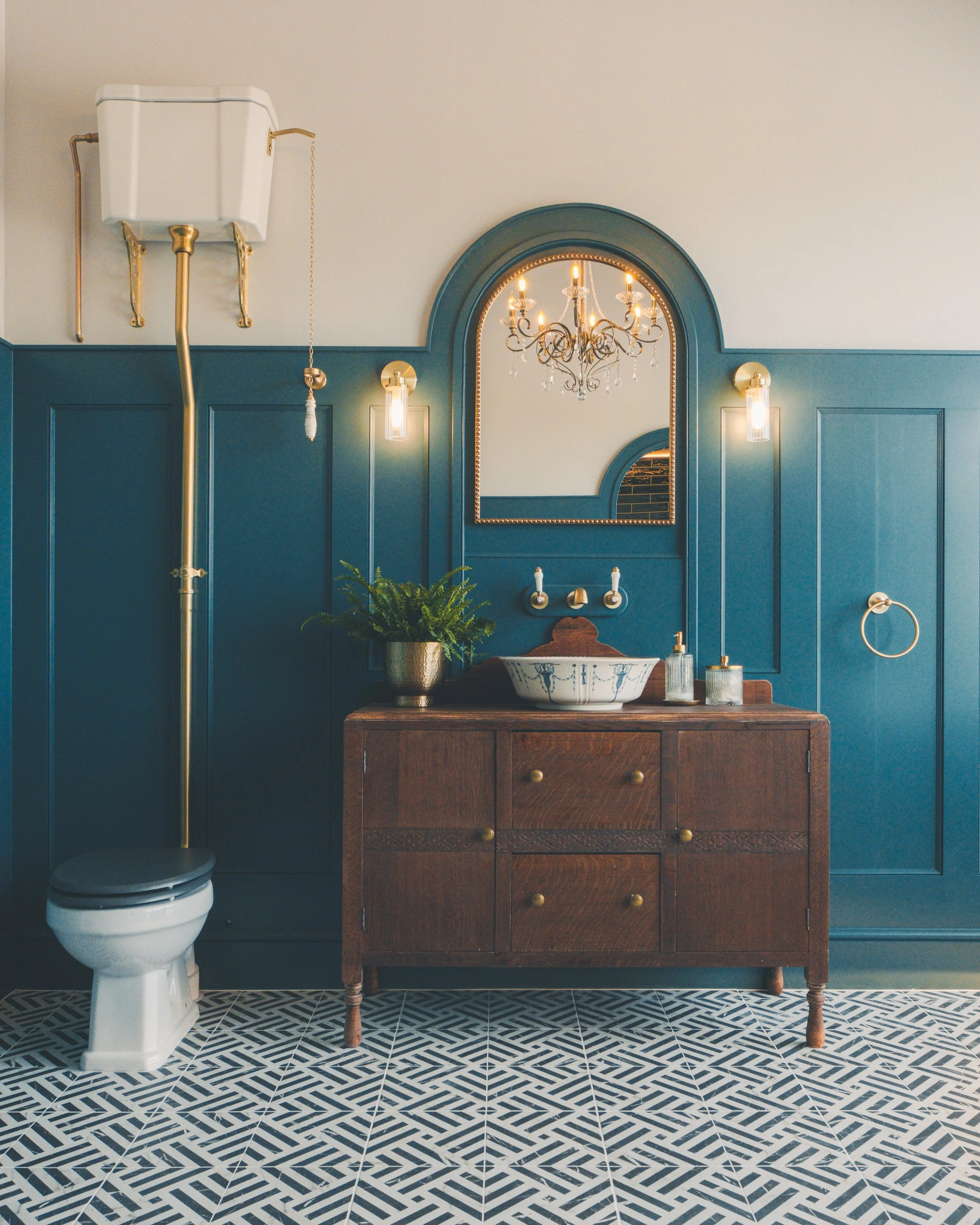A bathroom with a modern style, featuring a blue wall with paneling, a wooden vanity with brass knobs, a white vessel sink, a large arch-shaped mirror with a beaded frame, a chandelier with crystal accents, two wall sconces with glass shades, a potte