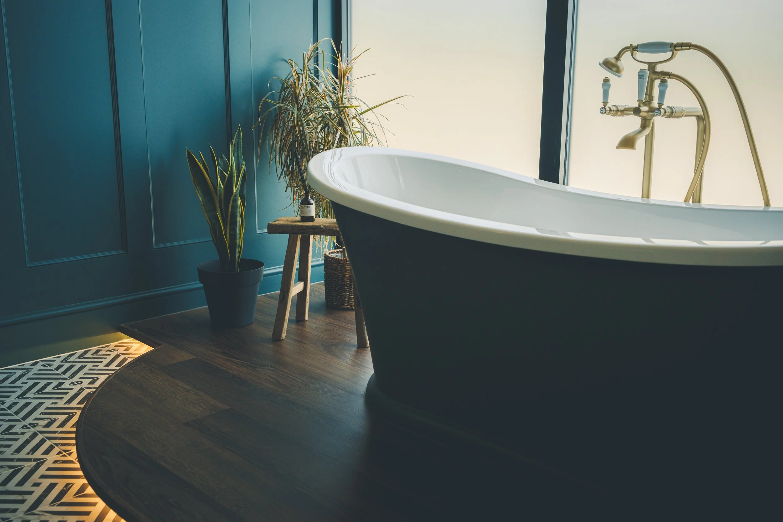 A modern bathroom featuring a white freestanding bathtub next to teal paneled walls, decorated with potted plants on a small wooden stool and a woven basket, illuminated by natural light from a window.