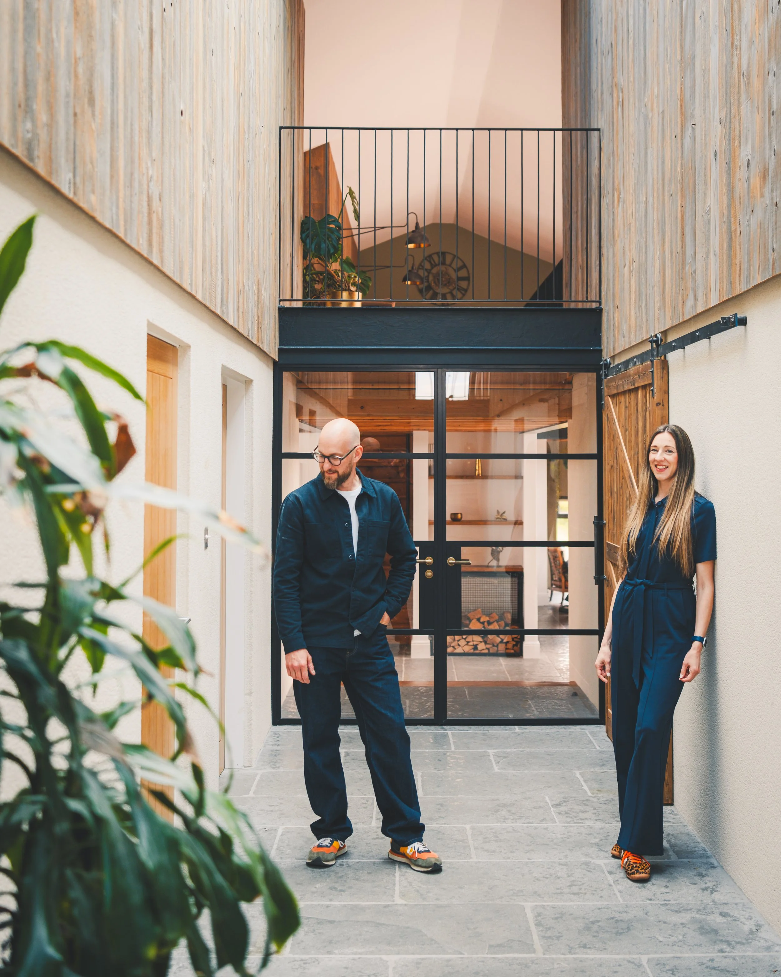Two people standing in a hallway with wooden and white walls, one man with glasses, bald head, and a beard wearing a dark blue jacket and colorful sneakers, and a woman with long hair wearing a dark blue jumpsuit and animal-print shoes, smiling.