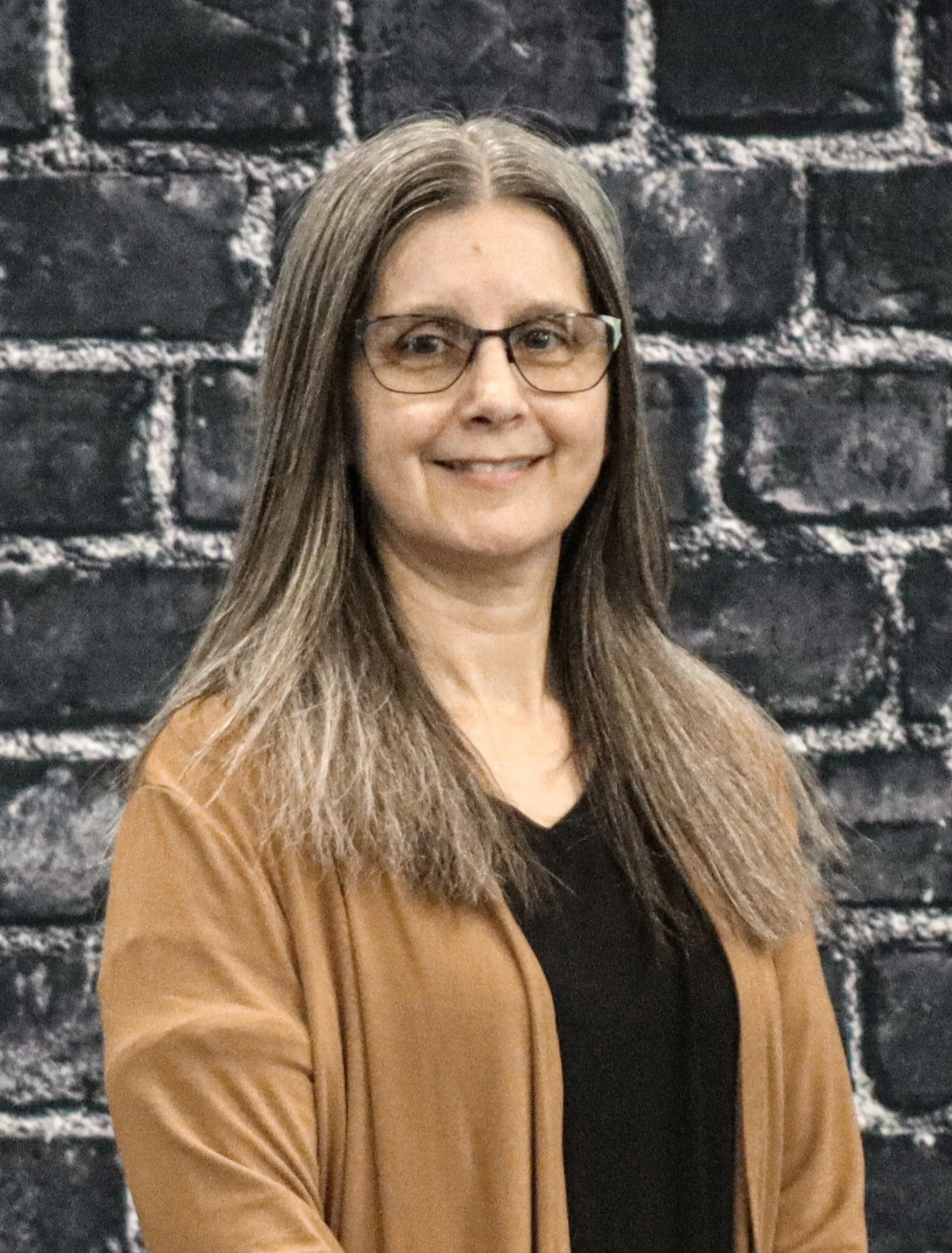 A woman with long brown hair and glasses smiling in front of a dark brick wall.
