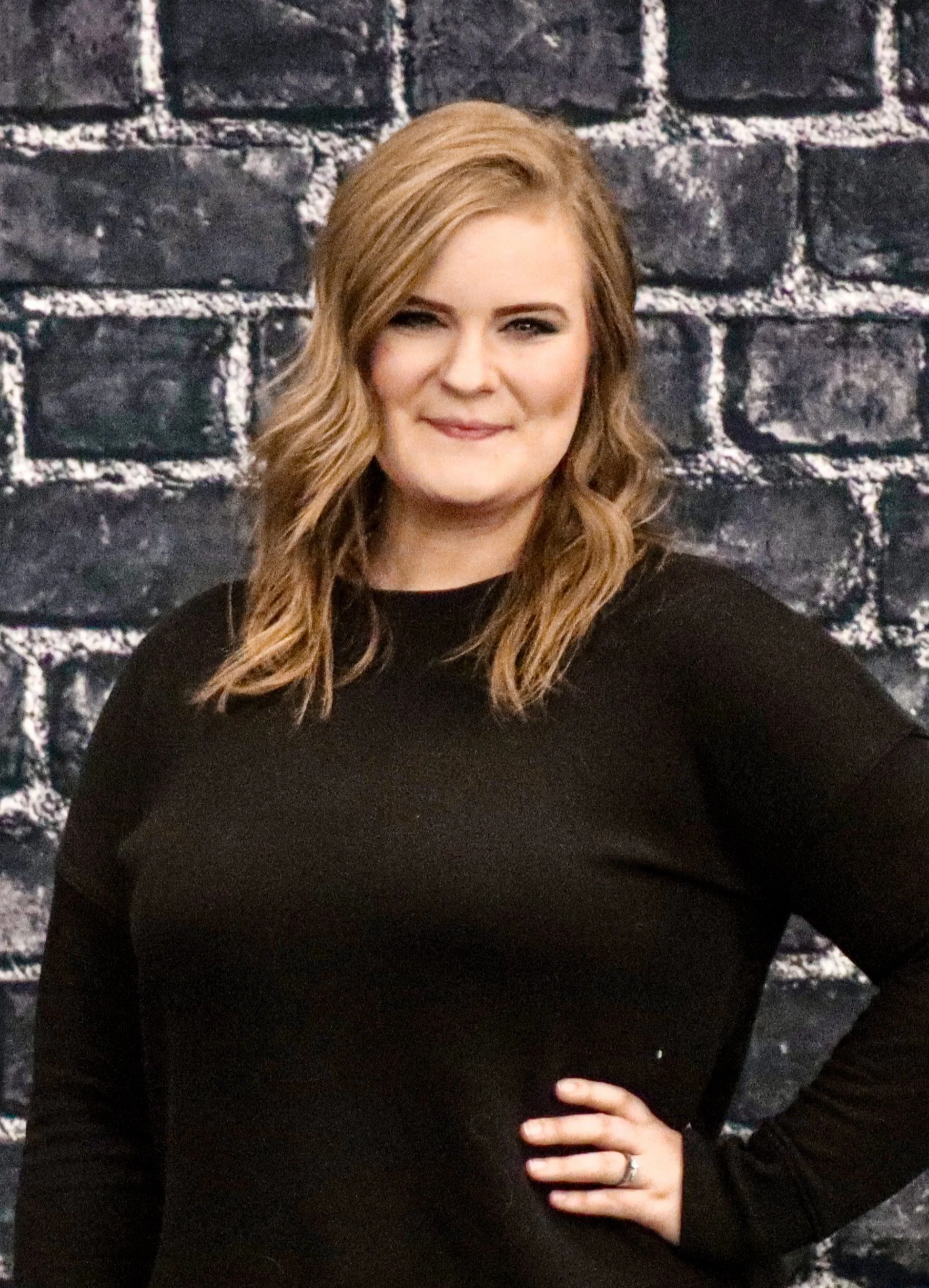 A woman with wavy, blonde hair, winking, wearing a black long-sleeved shirt, standing in front of a black brick wall.