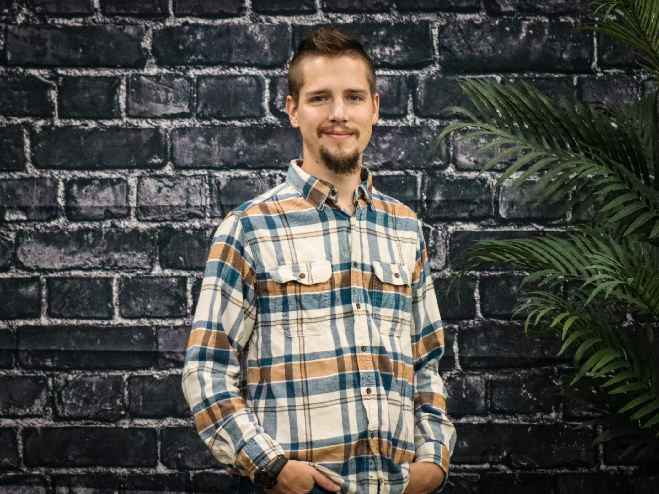 A young man with short brown hair and a goatee, wearing a plaid shirt, standing in front of a black brick wall with a green leafy plant to his right.