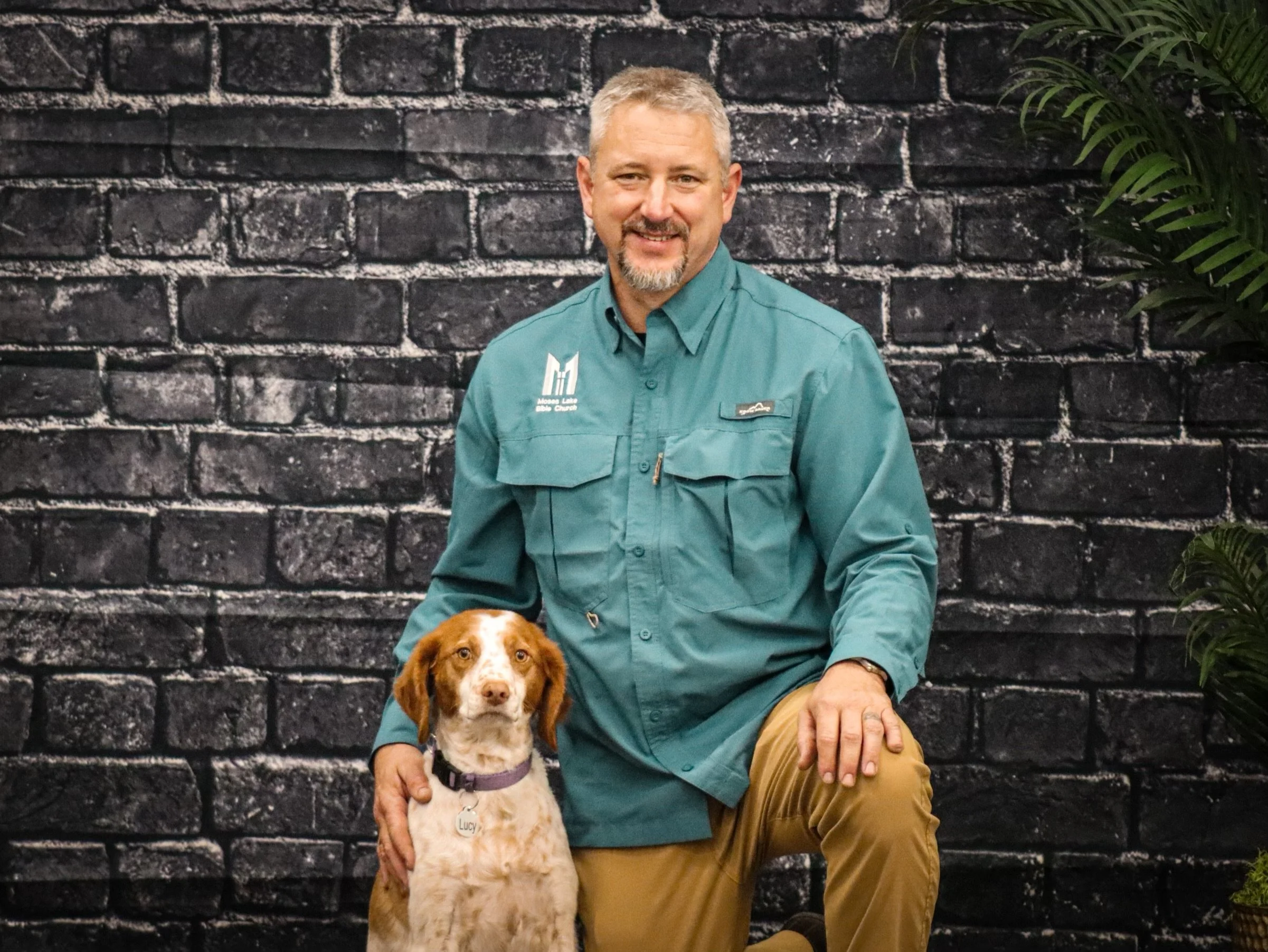 A man with short gray hair and a beard, wearing a teal shirt with a logo, is sitting against a dark brick wall with a green plant to the right. He has a brown dog with white markings, wearing a purple collar, on his lap.