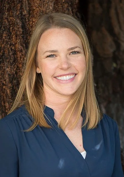 A young woman with blonde hair smiling, standing outdoors next to a tree.