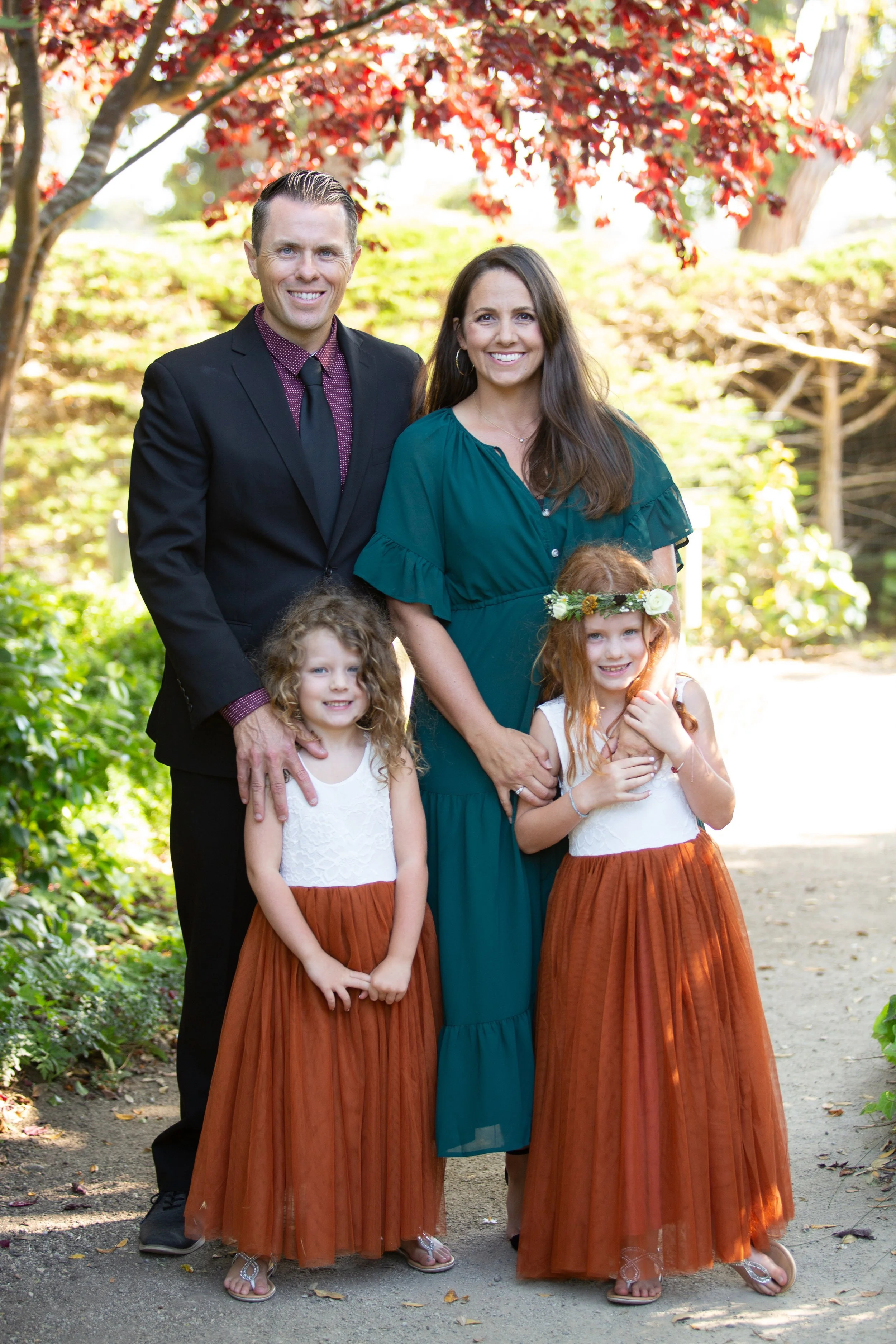 A family of five posing outdoors underneath a tree with red leaves. The father, dressed in a black suit, stands on the left. The mother, in a teal dress, stands in the middle. Two young girls with curly hair, wearing white tops and long orange skirts, stand in front. The girl on the right is wearing a flower crown.