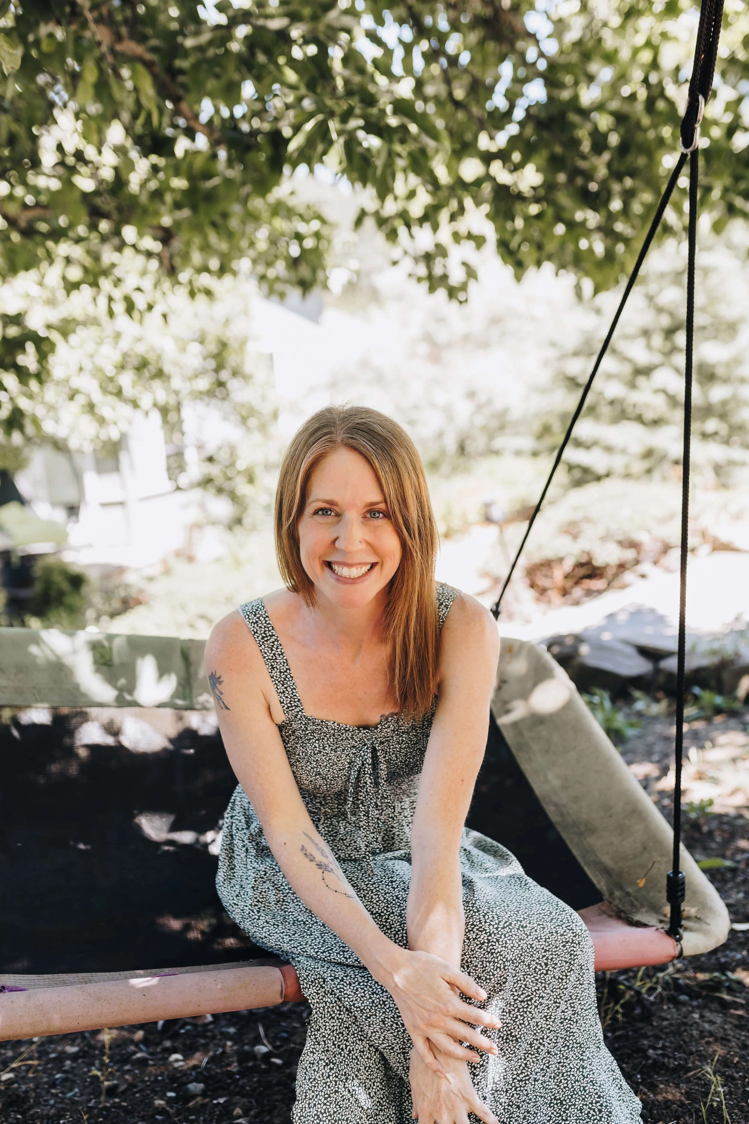 A woman with red hair and blue eyes smiling outdoors with green foliage in the background.