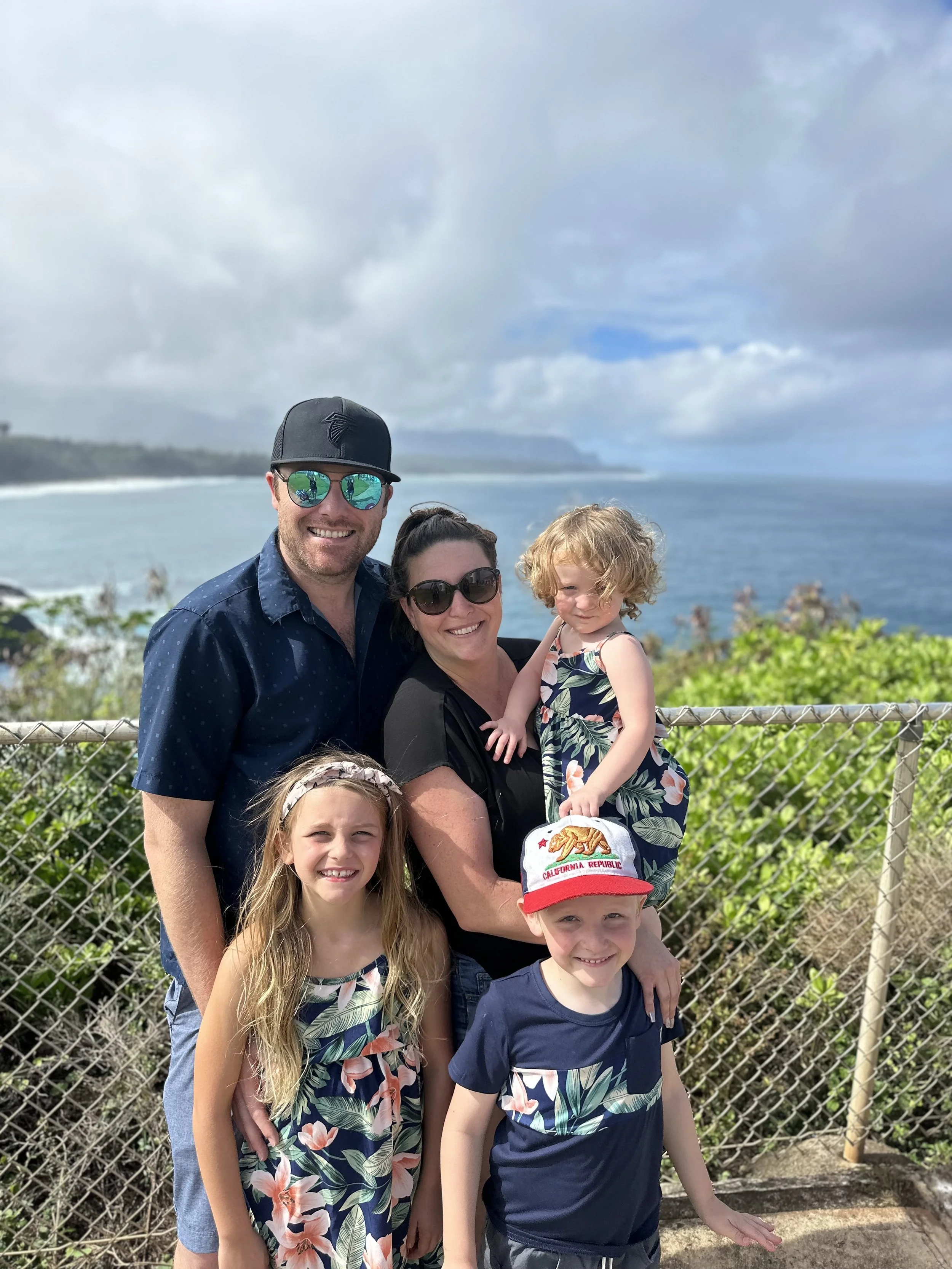 A family of six posing in front of a beach and ocean background with cloudy sky.