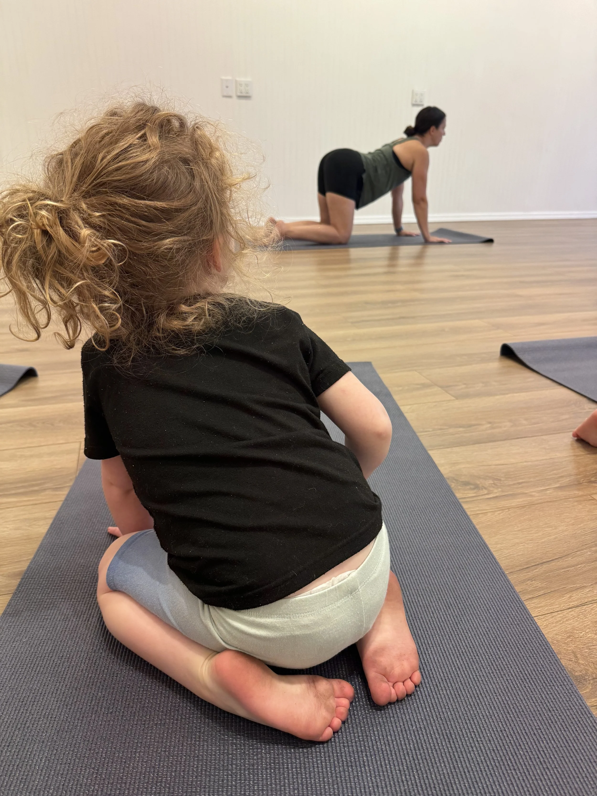A young girl with curly hair sitting cross-legged on a yoga mat, observing a woman in a yoga pose on a different mat in a spacious room with wooden floors and plain walls.