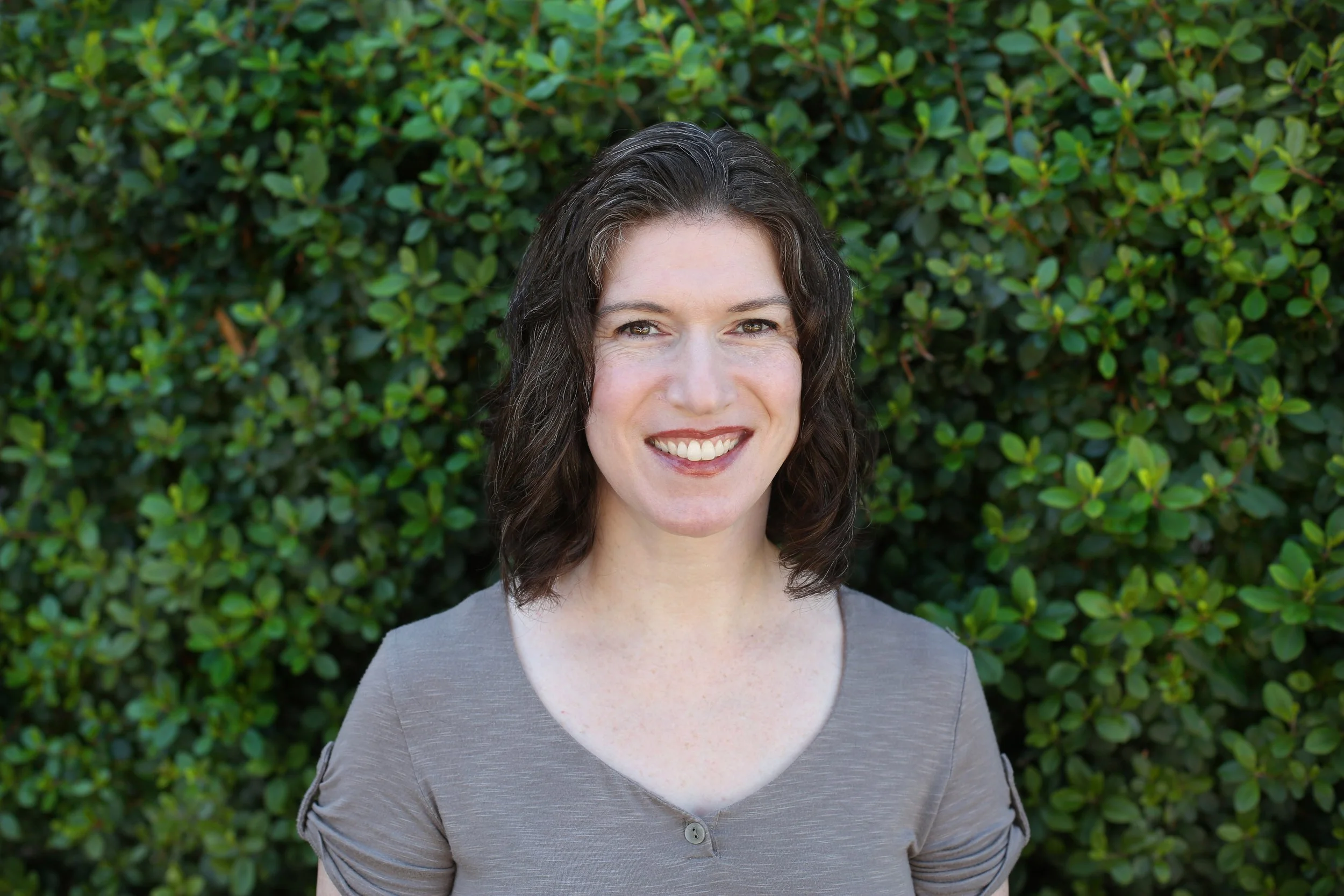 A woman with dark curly hair and light skin smiling in front of a green leafy bush, wearing a gray top.