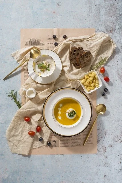 A flat lay of a breakfast setting on a table with a large bowl of yellow soup garnished with herbs, a small bowl of white sauce or yogurt with herbs, slices of dark bread, cherry tomatoes, blueberries, small cubes of cheese, and gold spoons, all placed on beige napkins and a newspaper.