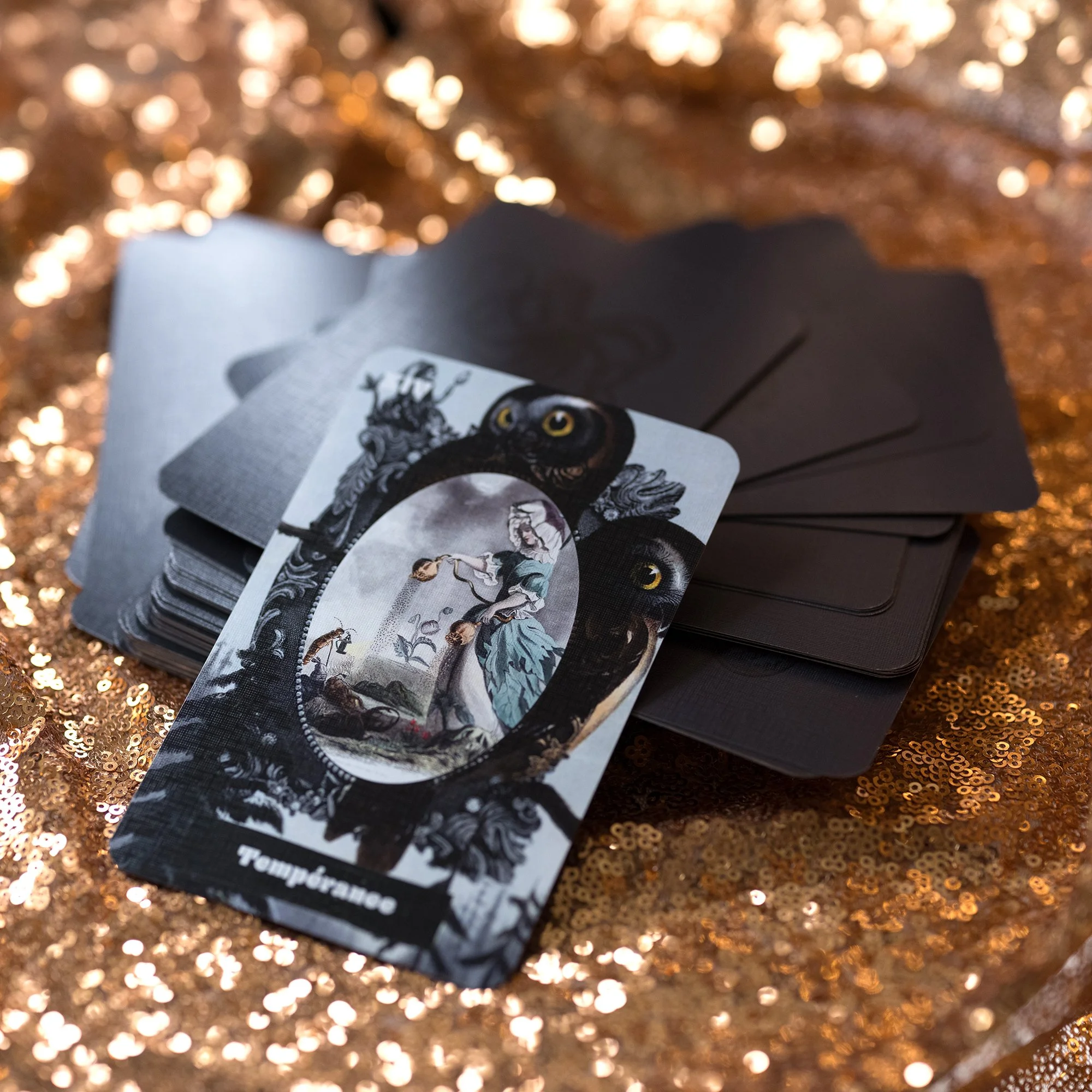 A deck of Tarot cards with one card labeled "Temperance" showing a woman pouring liquid from one container to another, with an owl and decorative black patterns on a shiny, gold, sequined surface.