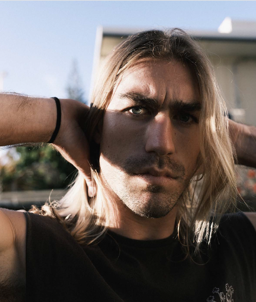 Close-up of a young man with long blonde hair, wearing a black shirt, outdoors on a sunny day, with sunlight partially casting a shadow on his face.