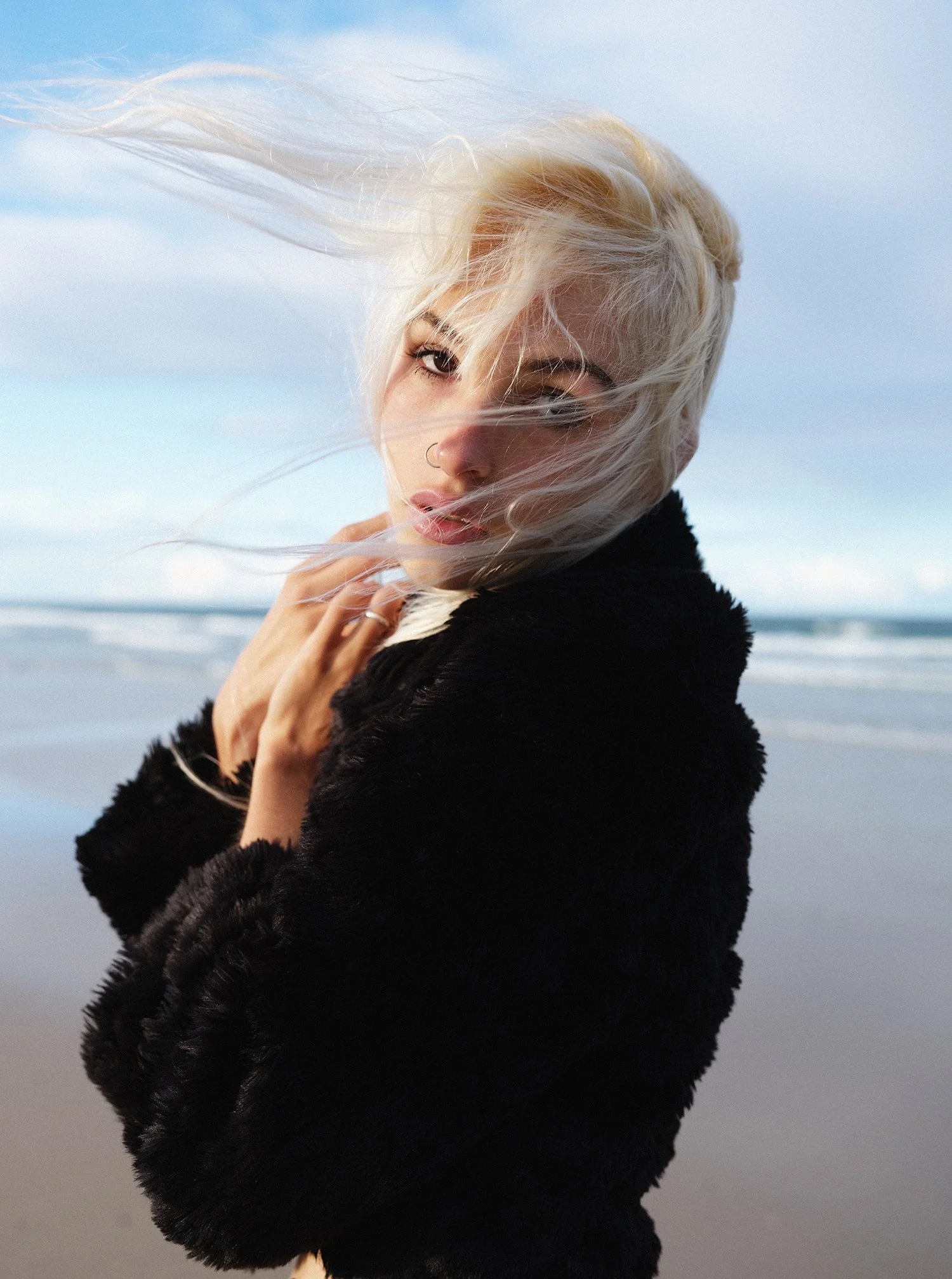 A woman with platinum blonde hair blowing in the wind at the beach, wearing a black fur coat.
