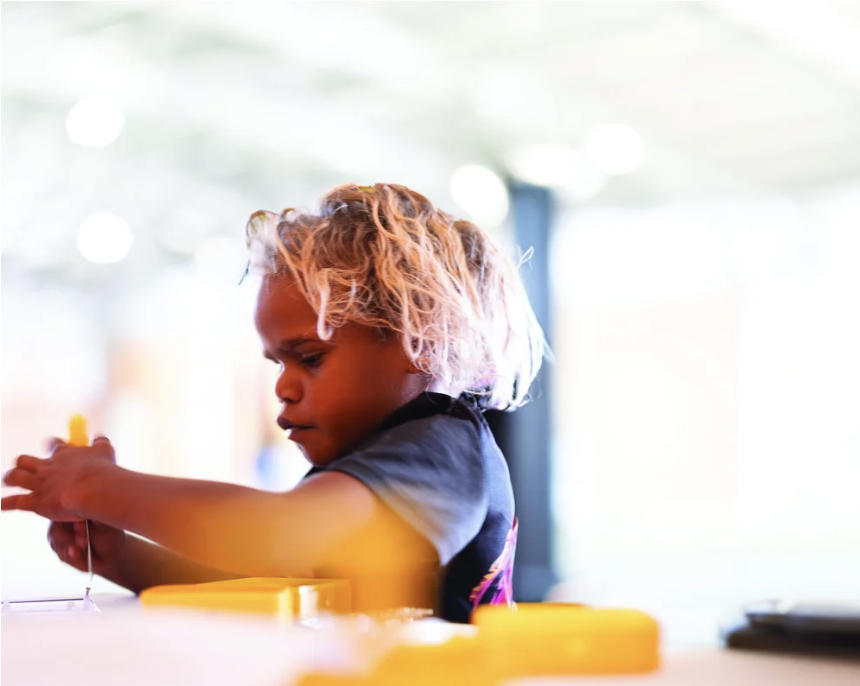 A young girl with curly blonde hair concentrating on a yellow object she is holding, with yellow items and a black device in the foreground on a white table.