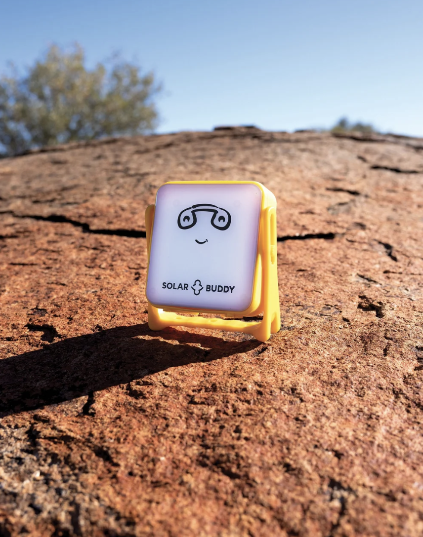 Solar Buddy device with a cartoon face, placed on a textured rock surface outdoors with blue sky and trees in the background.